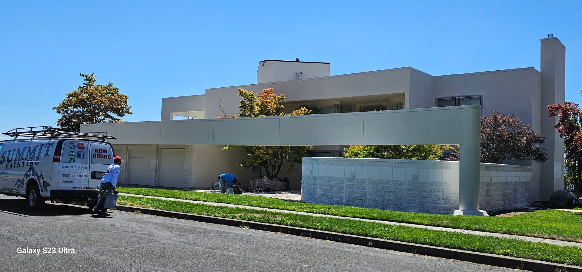 Modern light-colored house with a van parked in front. A person is working on the lawn on a sunny day.