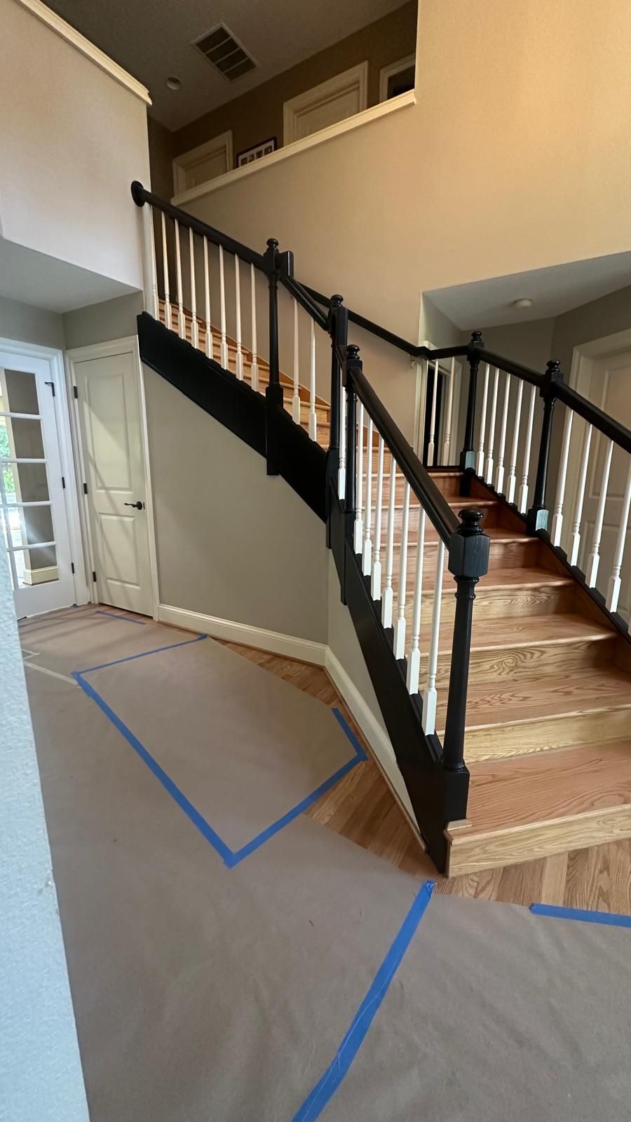 Staircase with black railing and white spindles; brown wooden steps. Protective paper and blue tape on floor.