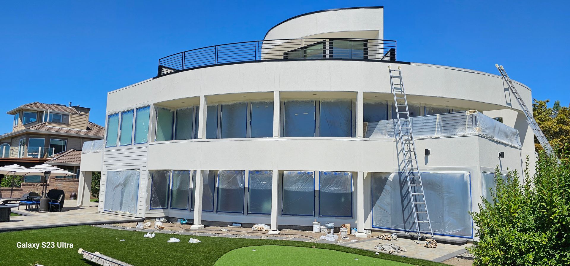 A large white, circular house under construction with scaffolding and ladders. Blue sky. Green lawn.