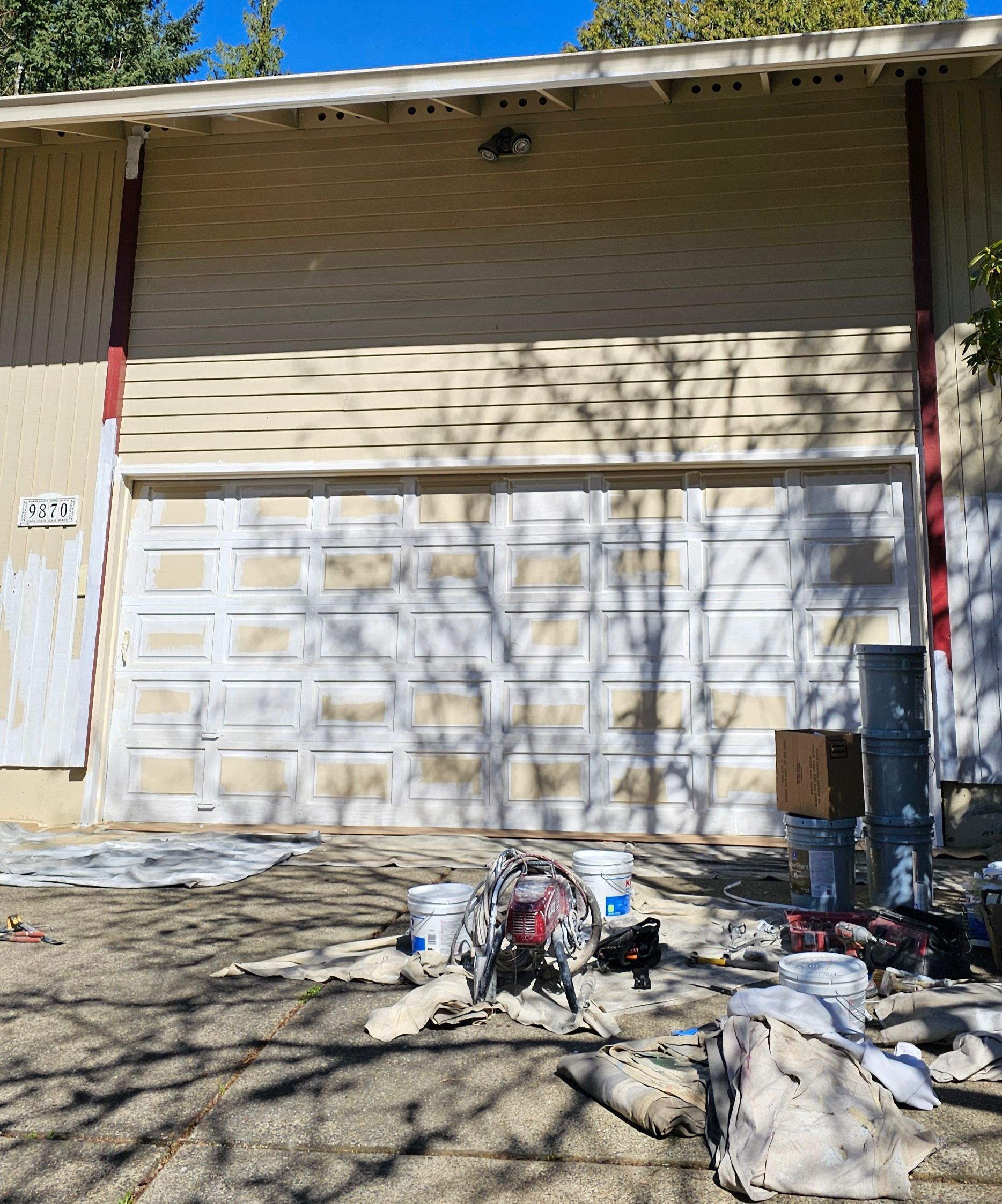 Garage door being painted white; painting supplies and drop cloths on driveway.