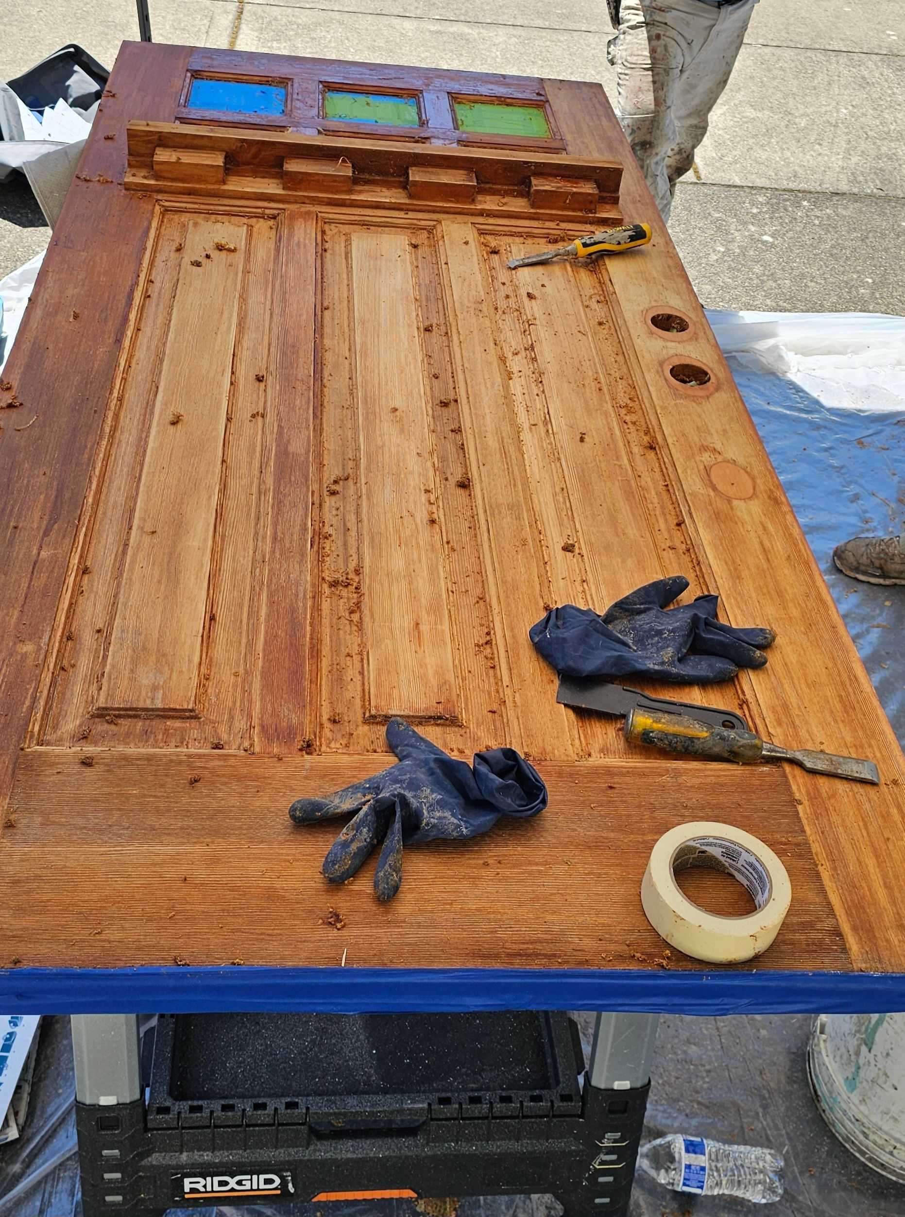 Wooden door being refinished on a table, with stained glass, tools, and gloves.