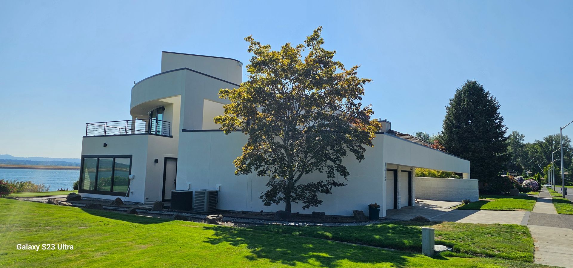 Modern white house with curved balconies, large windows, and a carport on a grassy waterfront lot.