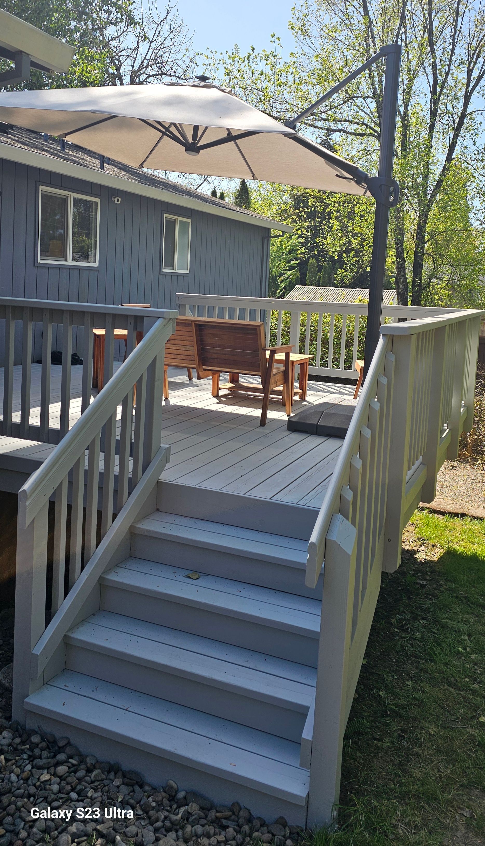 Gray deck with stairs leading up to it, a table, and an umbrella, next to a gray house.