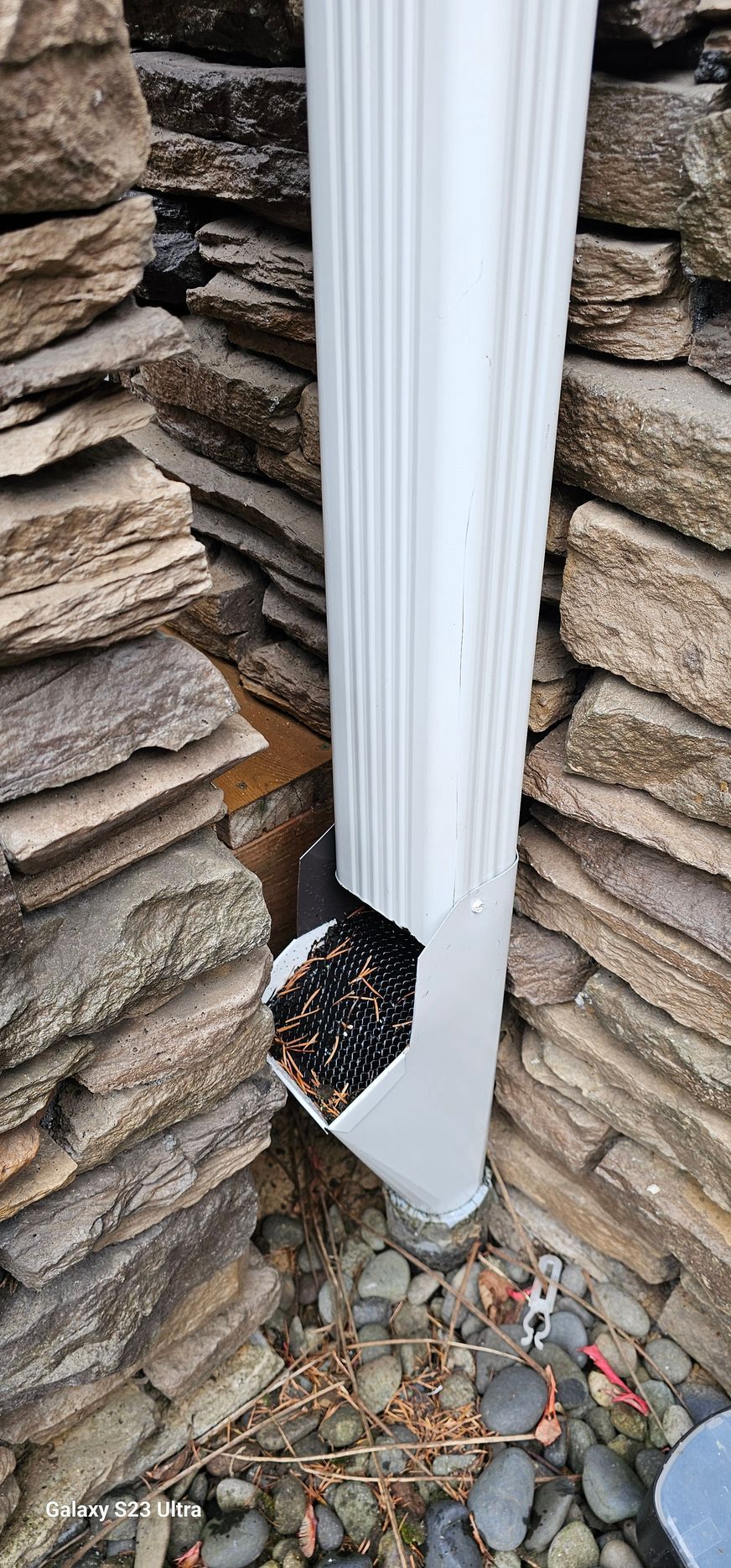 A white gutter filled with debris, nestled between two stone walls and gravel.