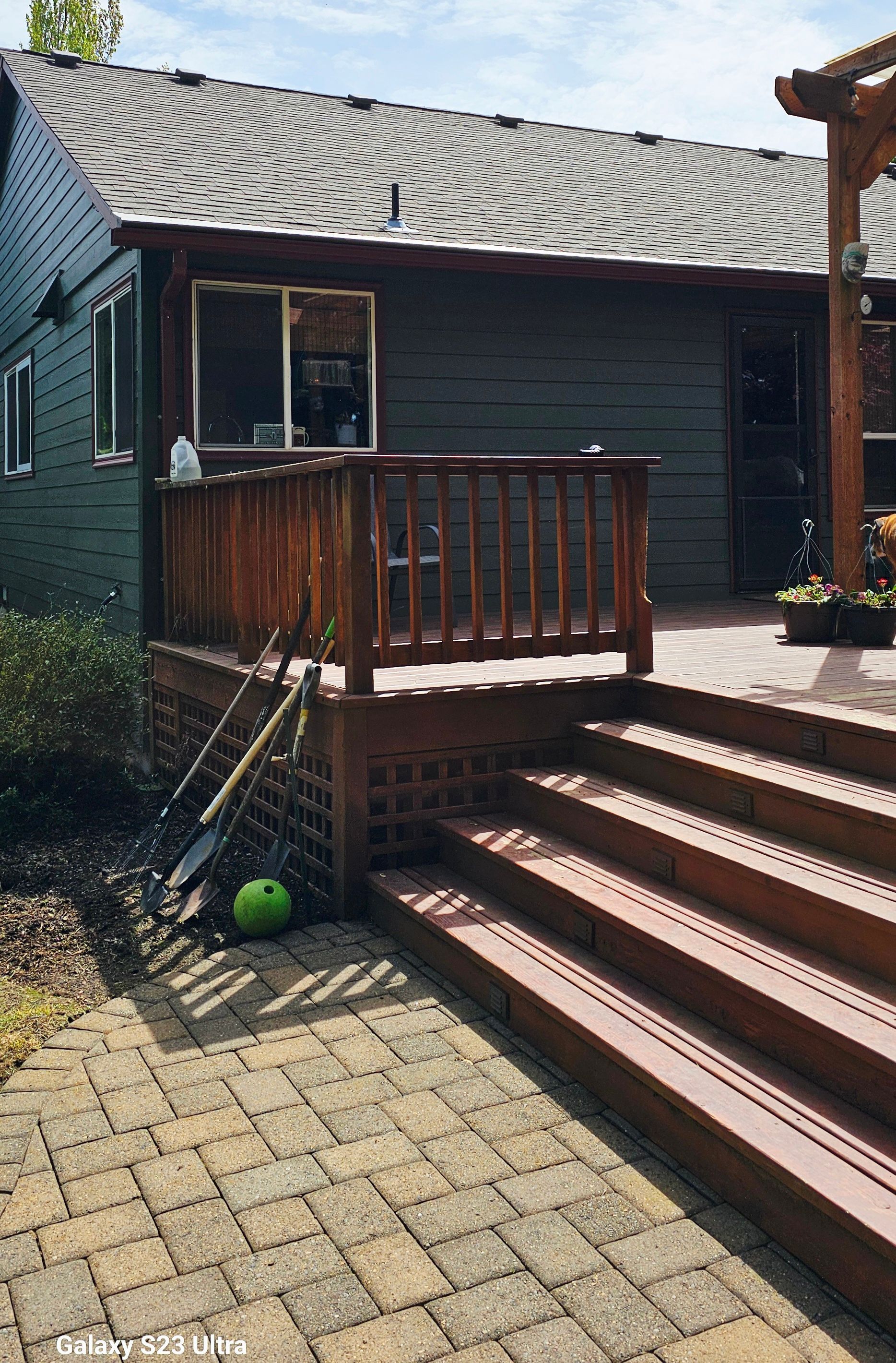 Backyard deck with wooden railing and steps leading to a brick patio. Tools and a green ball are leaning against the deck.