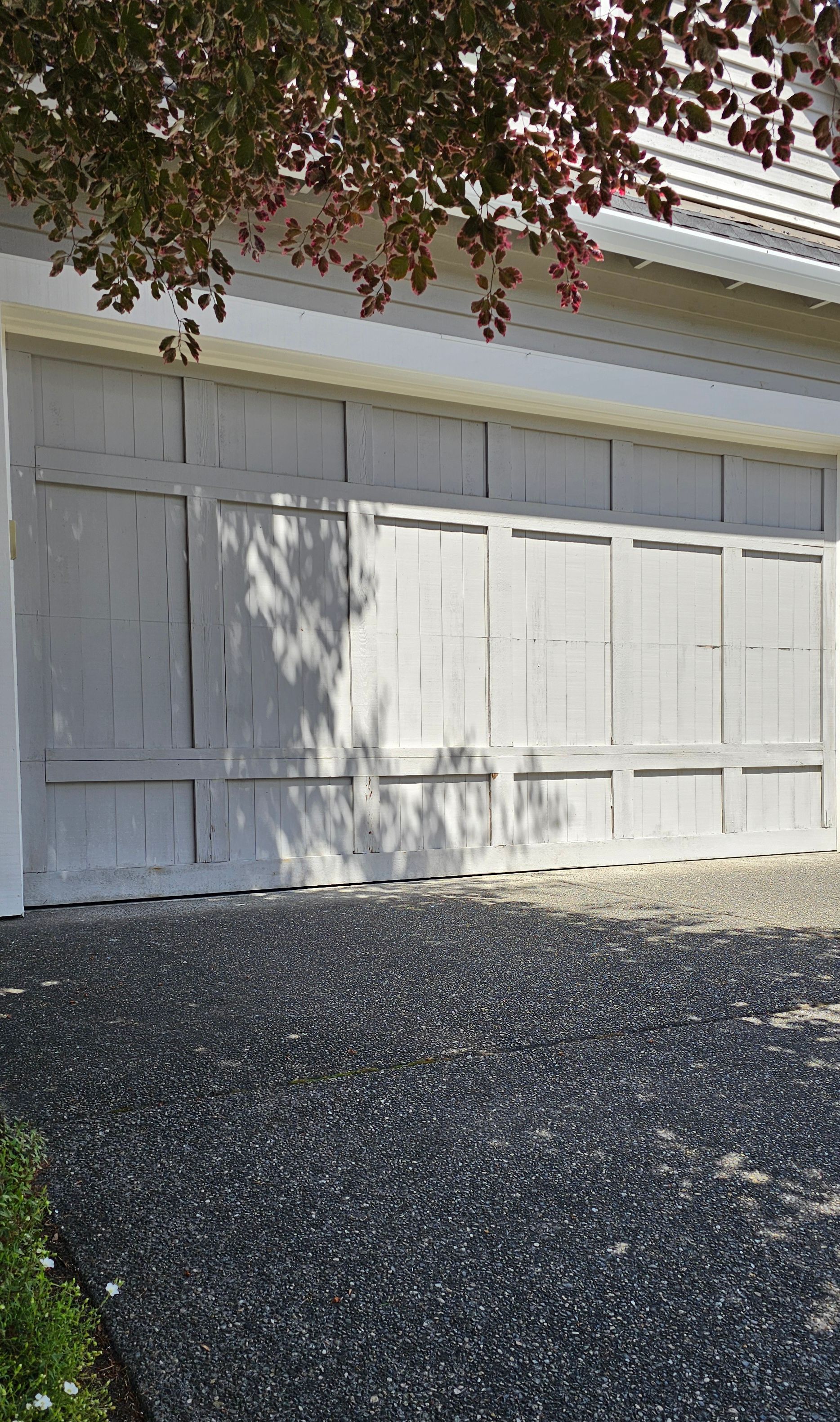 A light-colored garage door with a gravel driveway and tree above.