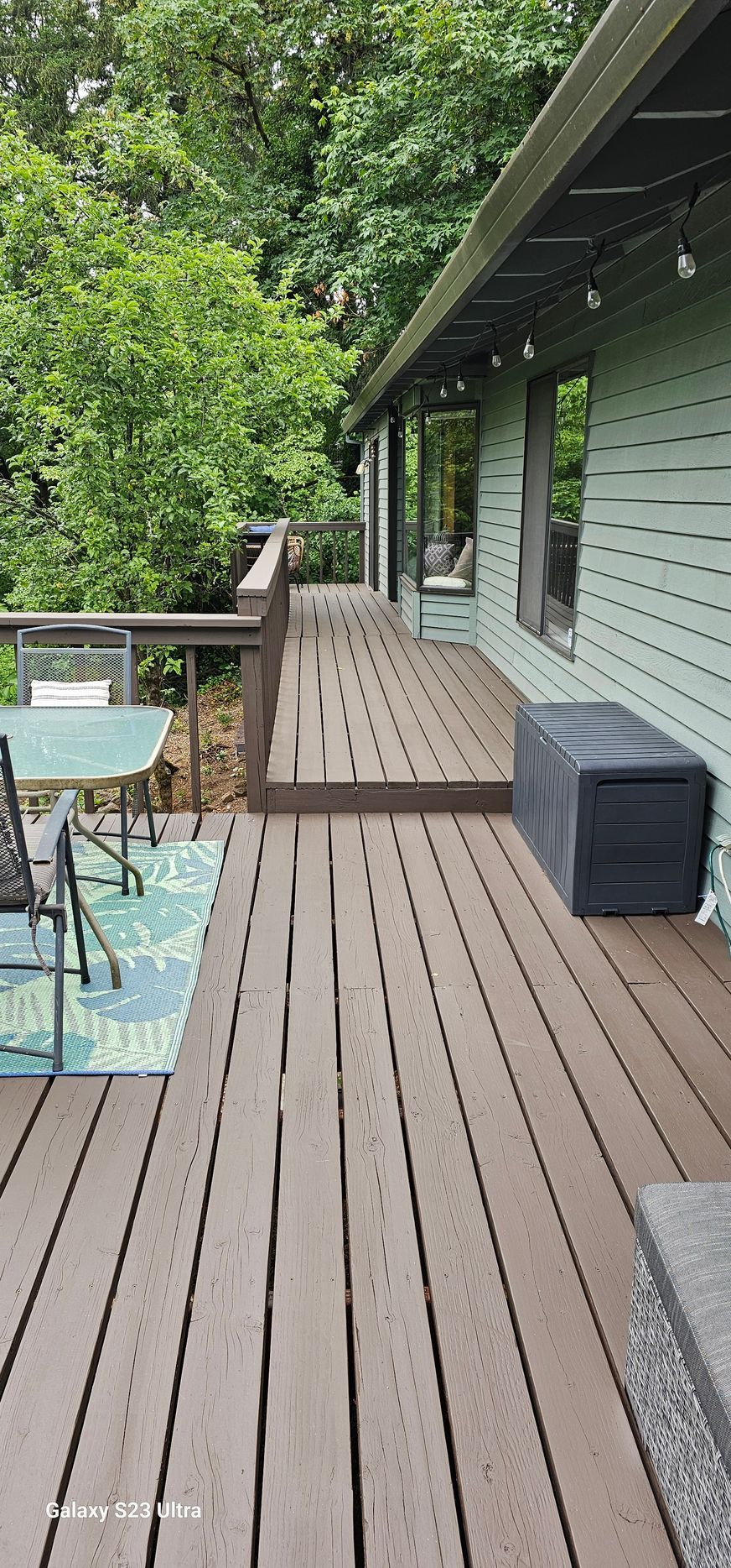 Wooden deck with table and chairs. Green foliage and house siding.