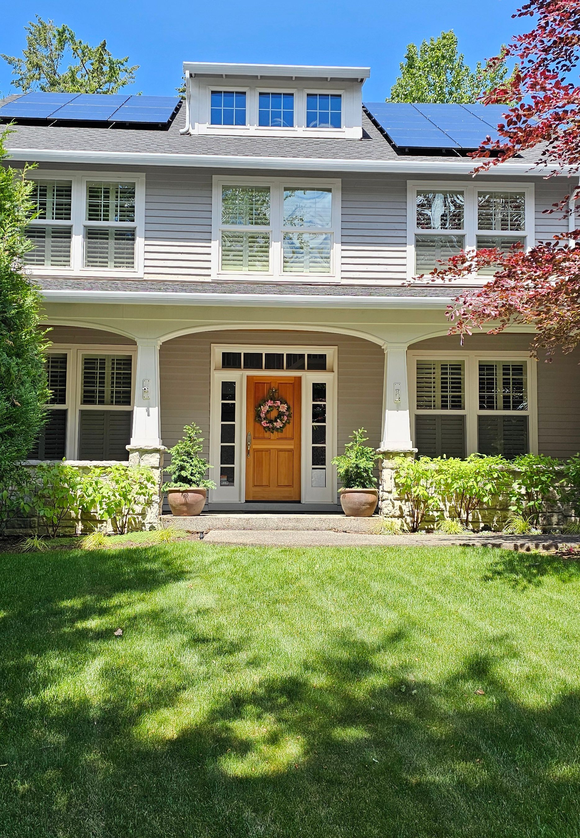 Two-story house with beige siding, a wooden front door, porch, and solar panels on the roof. Sunny day.