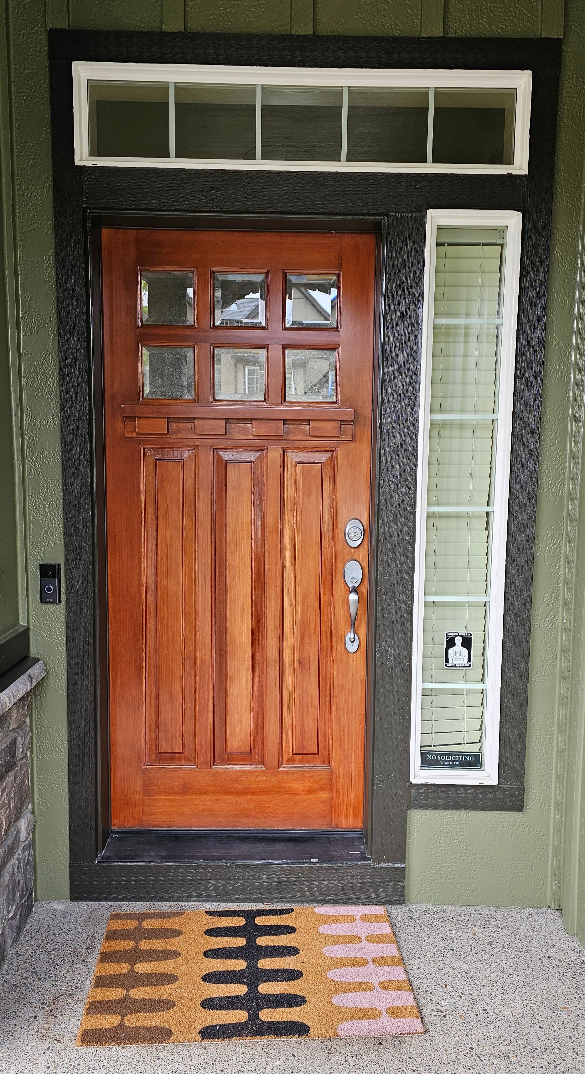 Wooden front door with sidelight, surrounded by black trim, on a green exterior. A patterned doormat is in front.