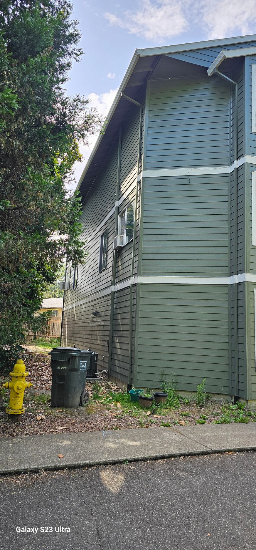 Side view of a two-story green building with a yellow fire hydrant and a trash bin in front.