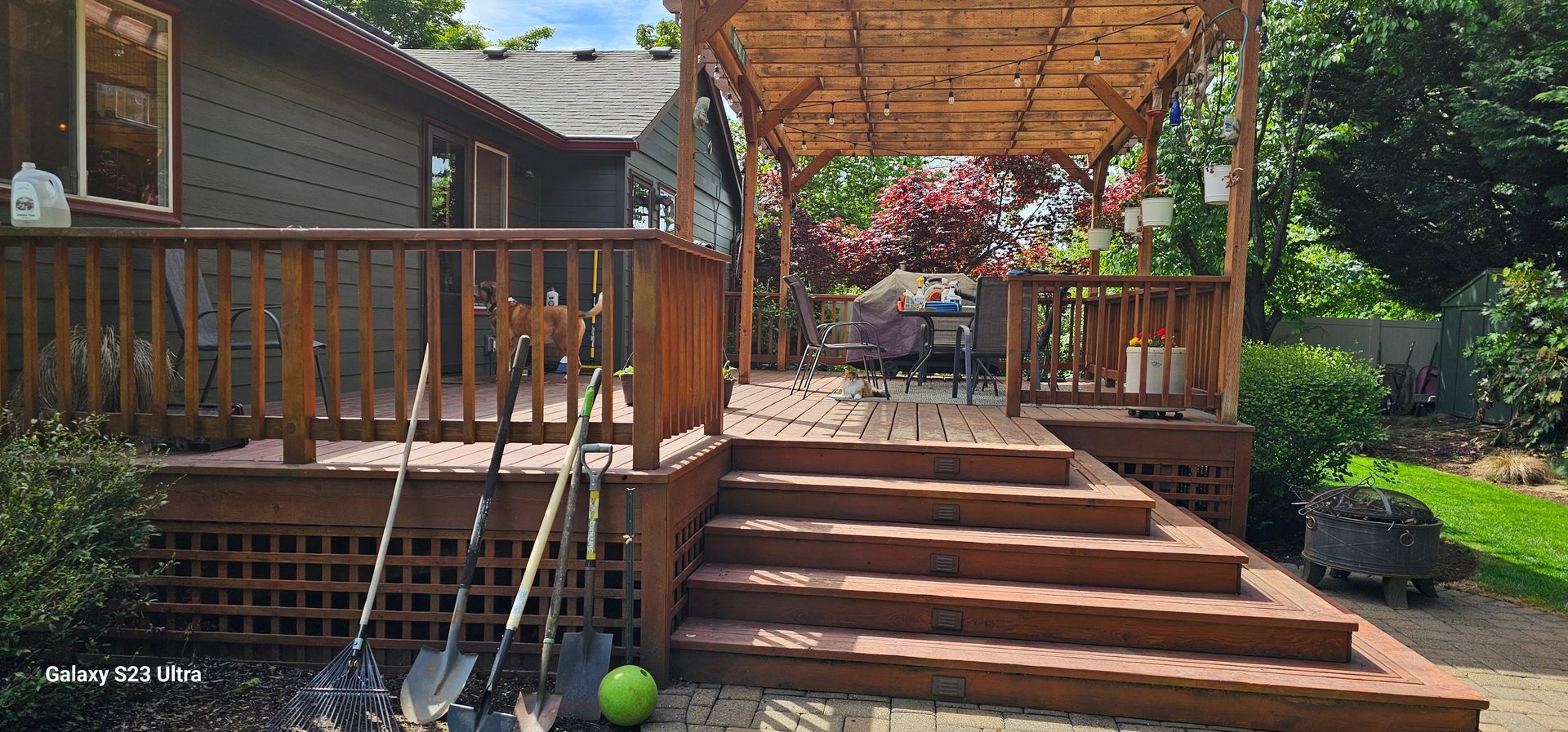 Wooden deck with steps leading to a shaded seating area. Shovels are leaning against the railing. Lush greenery surrounds the space.