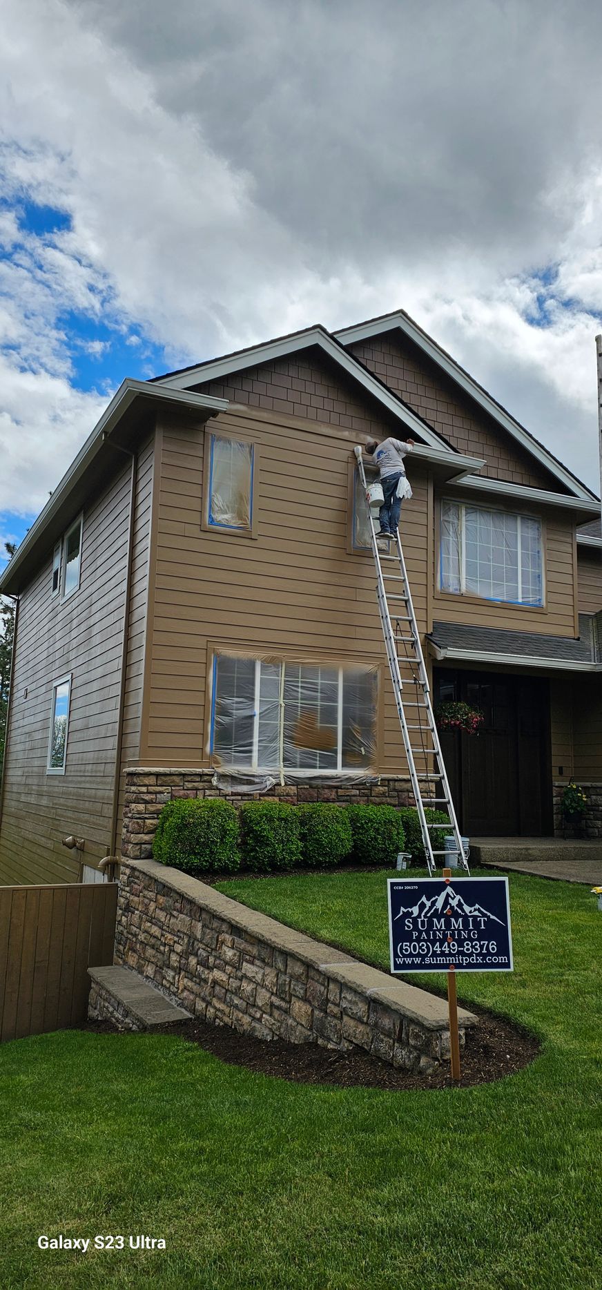 A person on a ladder is working on the siding of a two-story house.