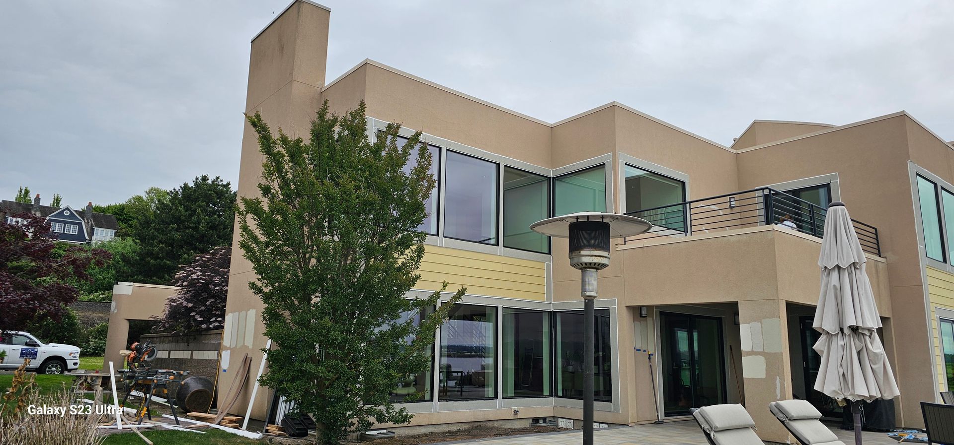 Modern, two-story beige house with large windows, patio, and partially obscured by a tree. Overcast sky.