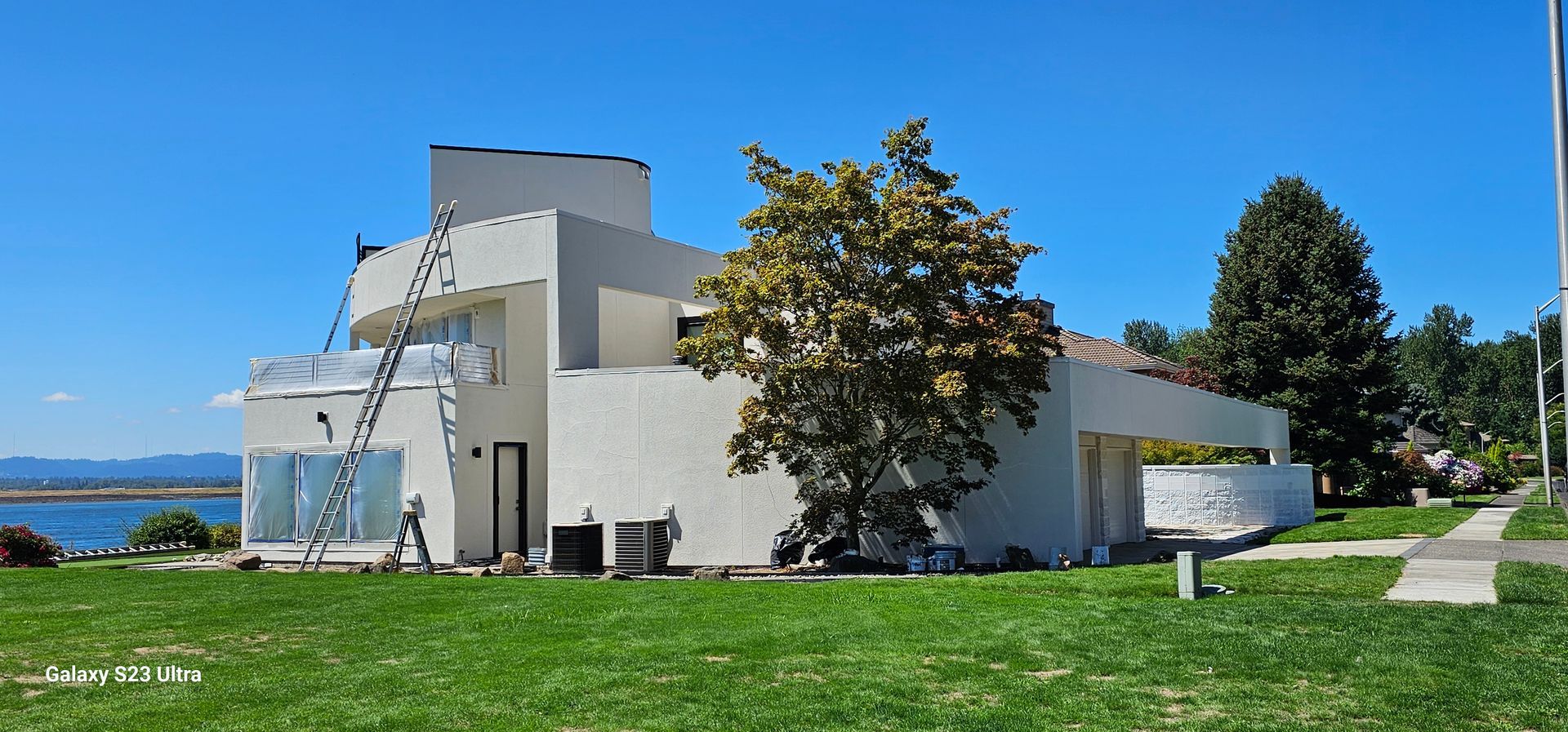 White modern house with a ladder, green lawn, trees, and a view of the water under a clear blue sky.