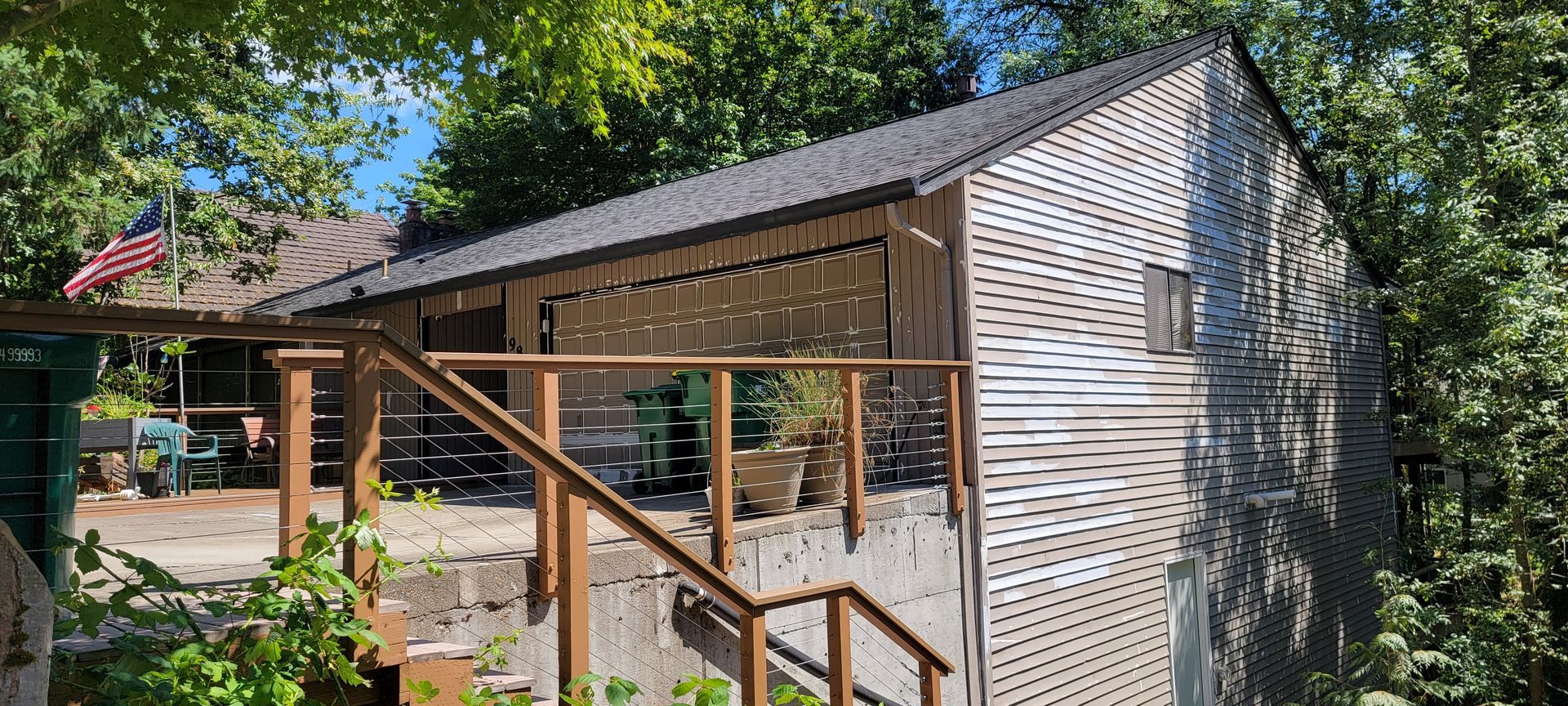 Wooden house with slatted facade, deck with railing, stairs, and American flag. Surrounded by trees.