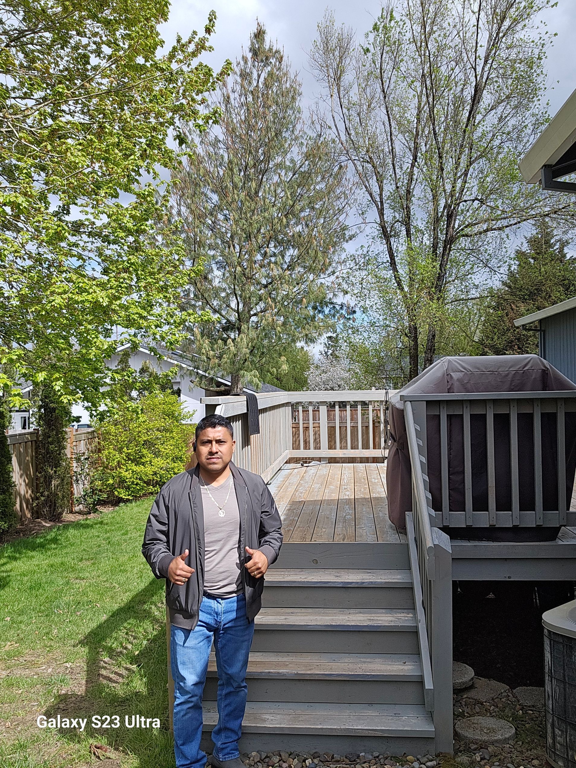 Man in gray jacket and jeans stands on wooden steps outside, trees and blue sky background.
