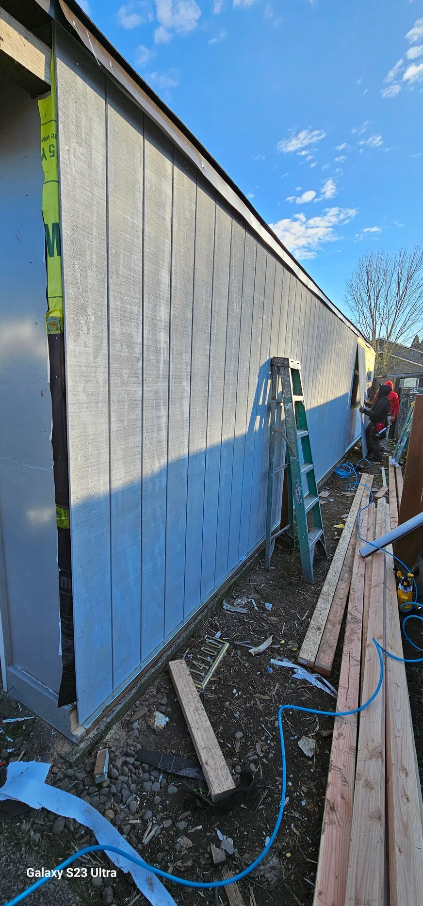 Construction in progress: siding installation on a building's exterior with a ladder, wood planks, and blue sky.