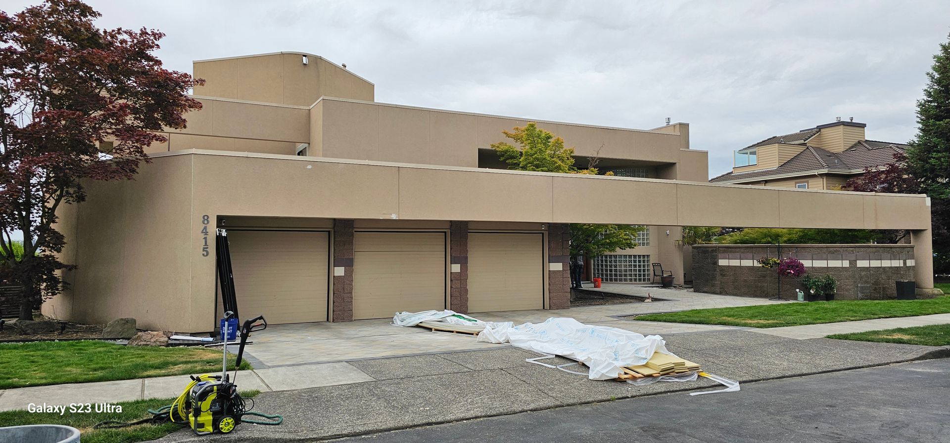 A beige two-story house with three garage doors and a pressure washer in the driveway.