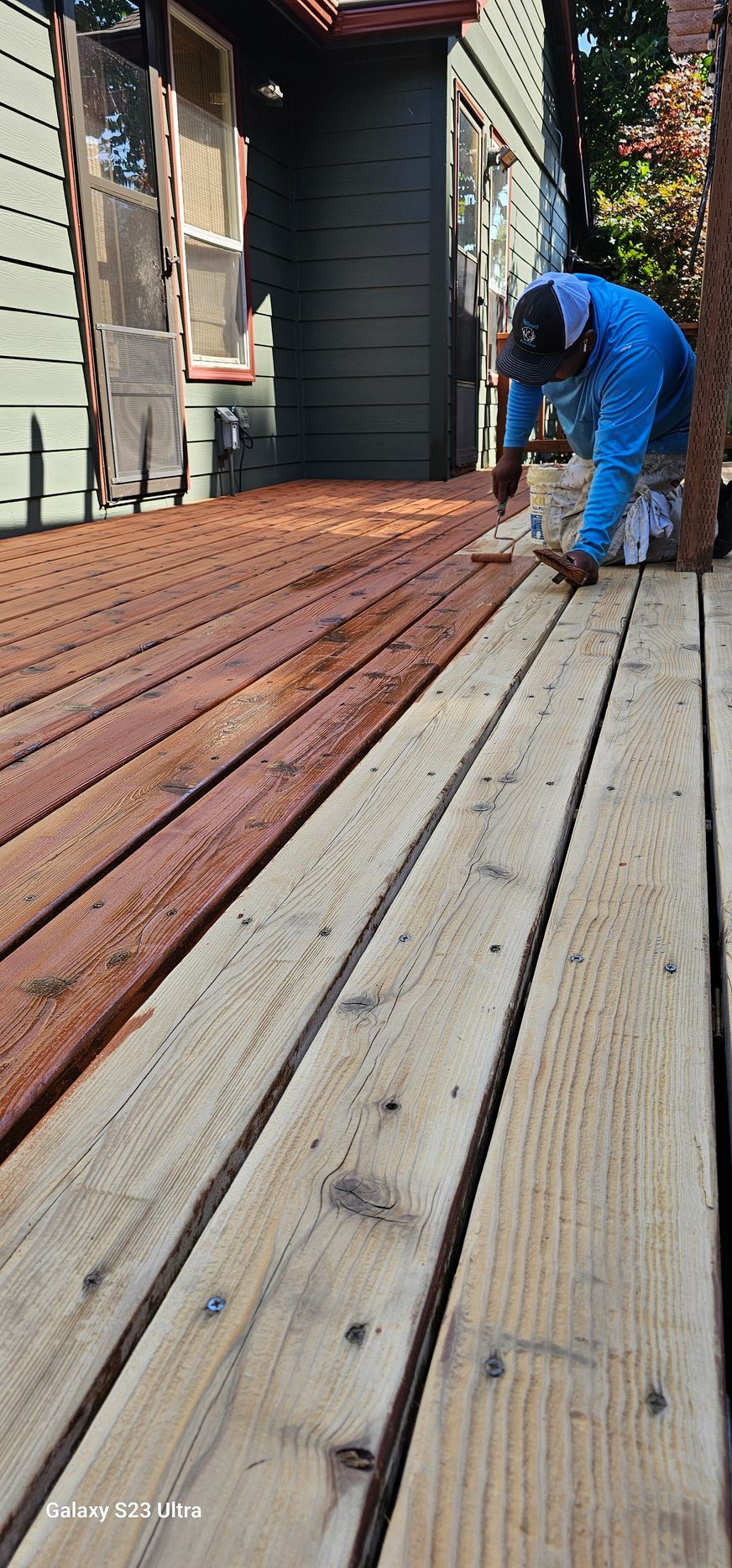 A person paints a wooden deck with reddish-brown stain; some boards are stained and some are not.