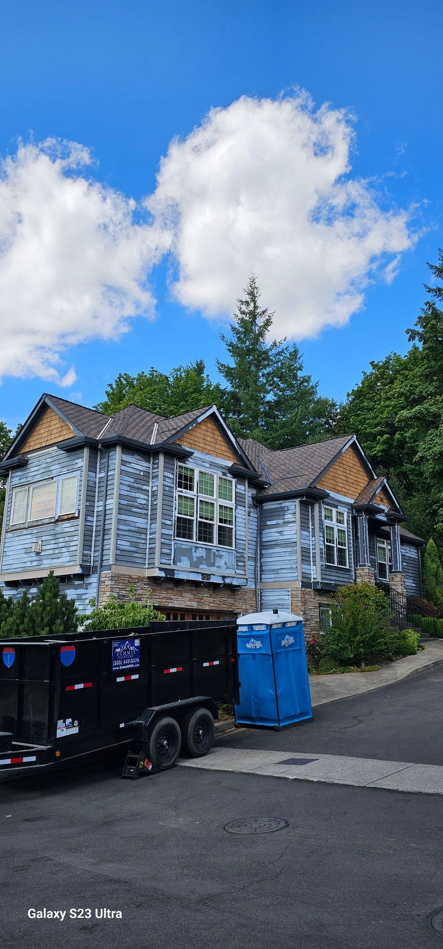 House under construction with exposed walls, a trailer, and portable toilet on a sunny day.