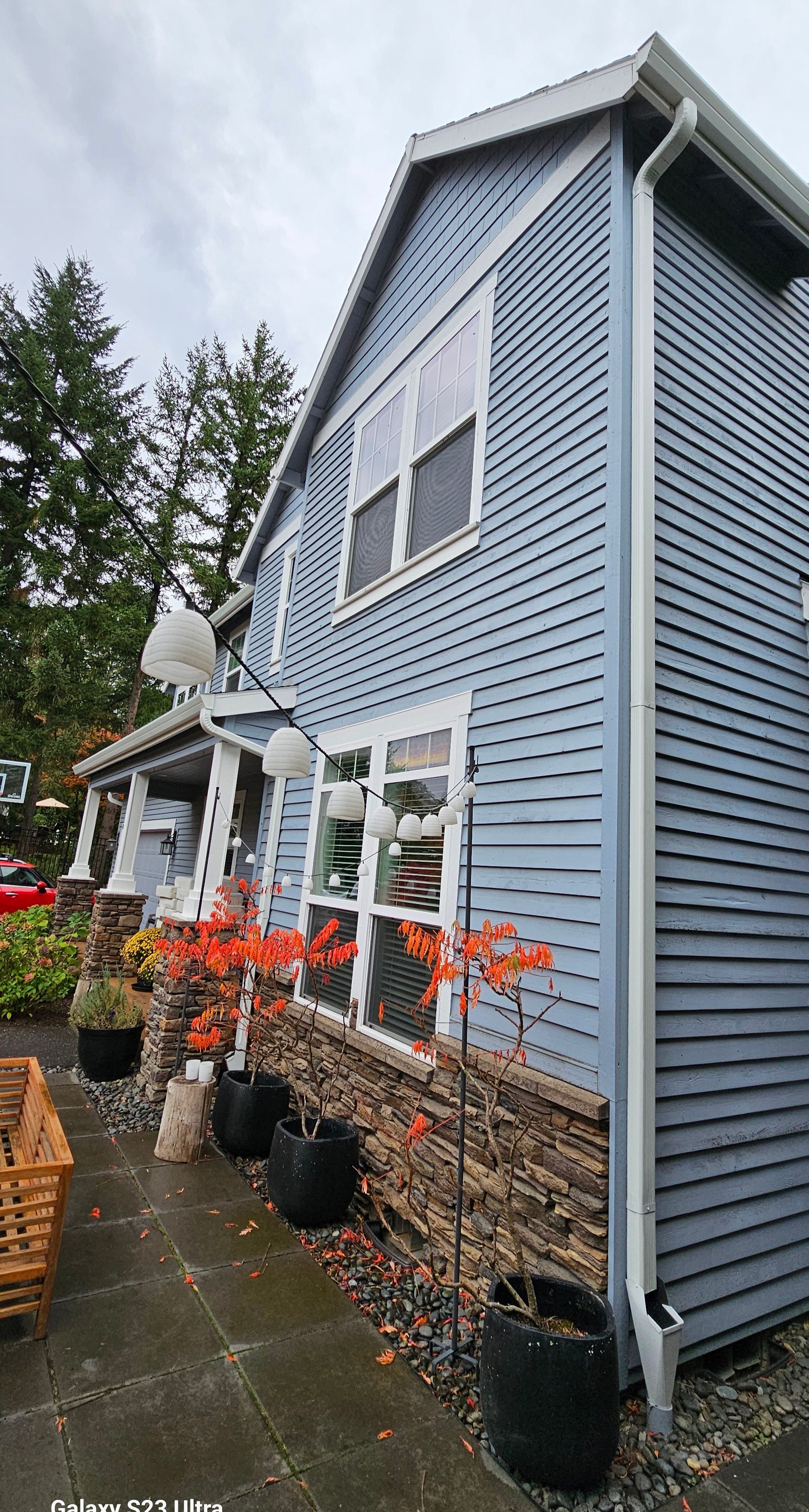 Blue house with white trim, brown stone facade, plants in pots and autumn foliage in front.