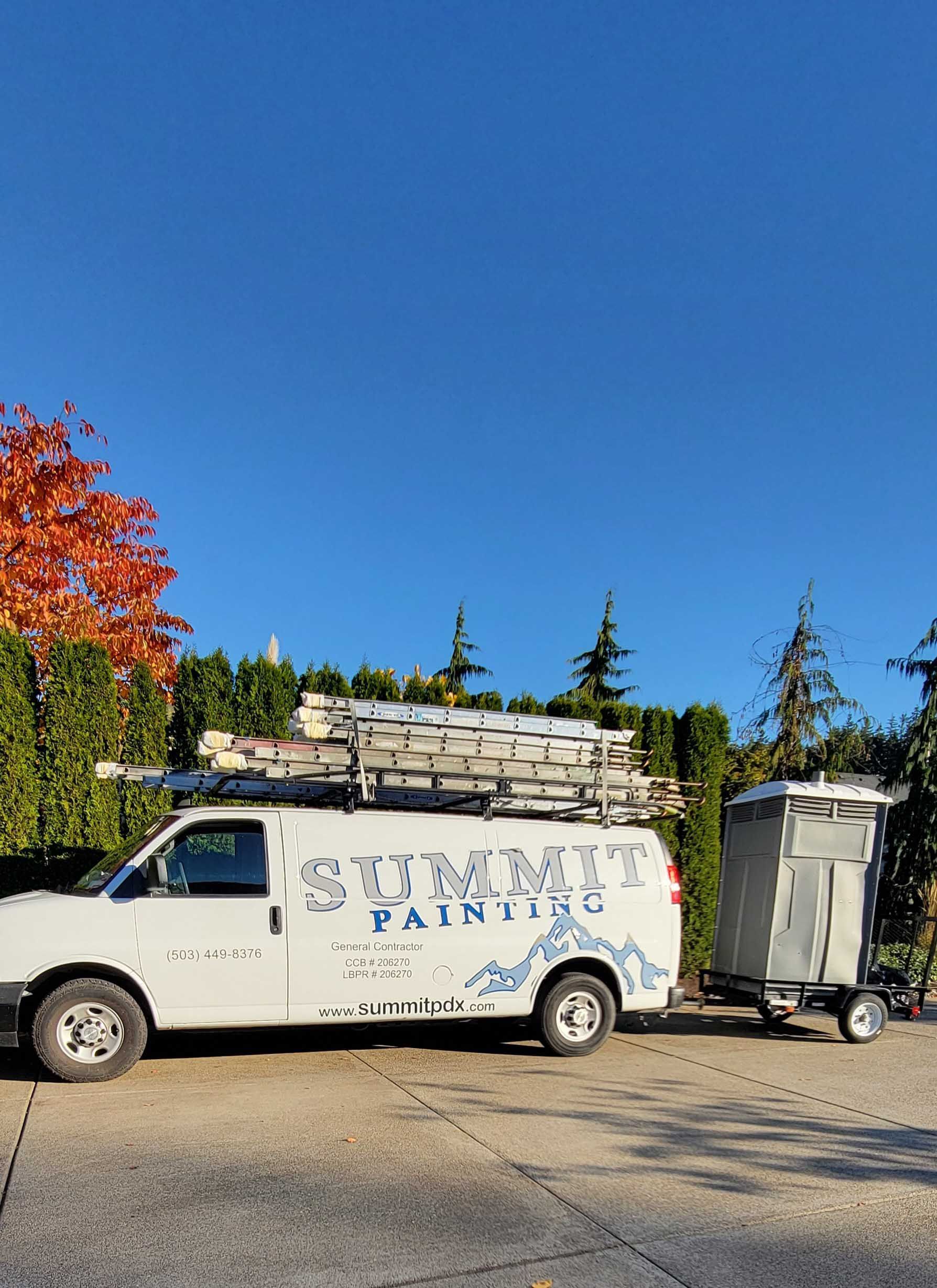 White Summit Painting van with ladders on top, towing a portable toilet. Autumn leaves and blue sky.