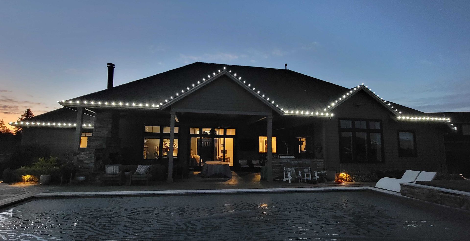House exterior at dusk with white lights outlining the roof, reflecting in the pool.