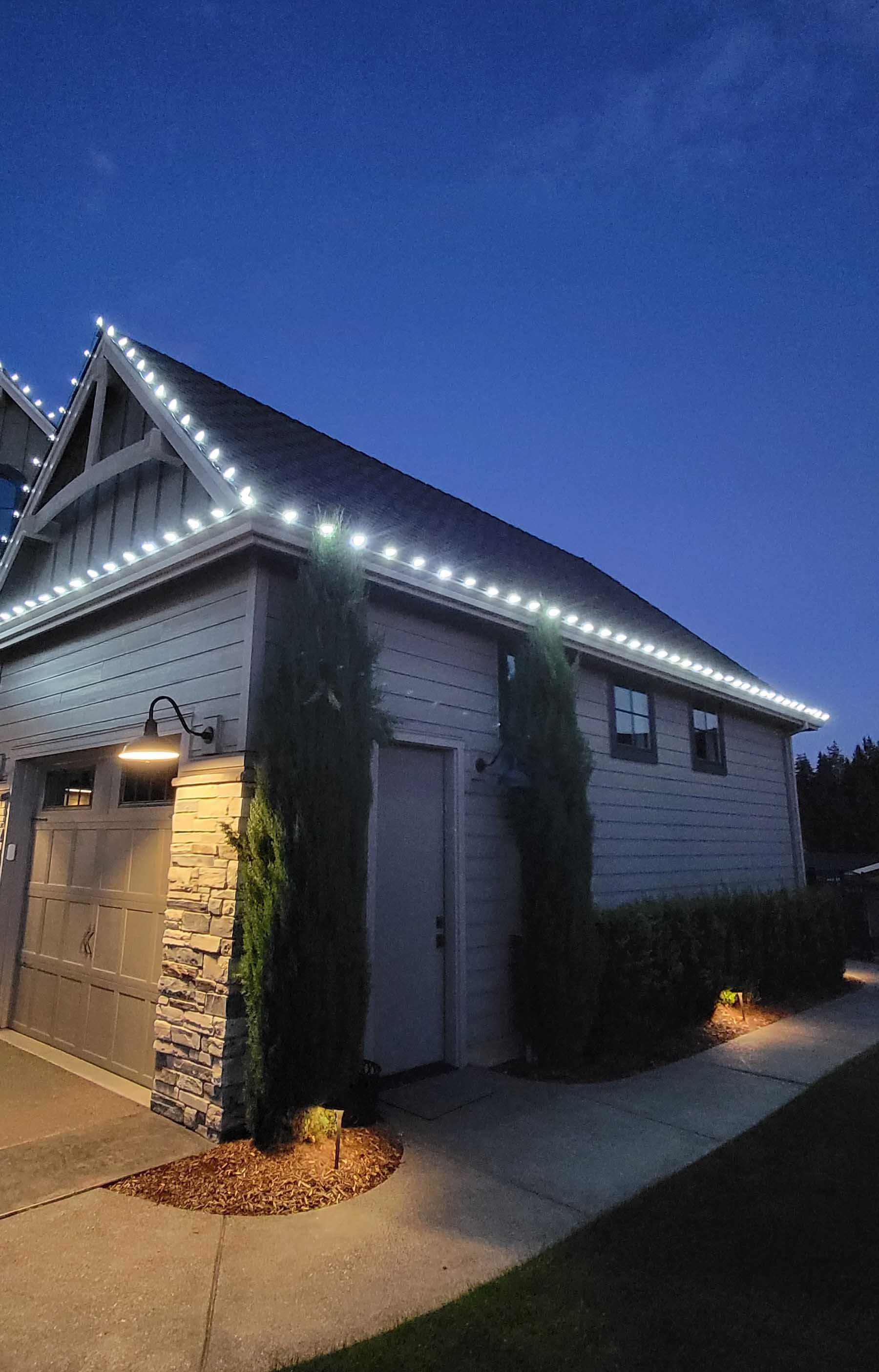 Garage with white Christmas lights on the roof at dusk. Two tall green trees stand in front.