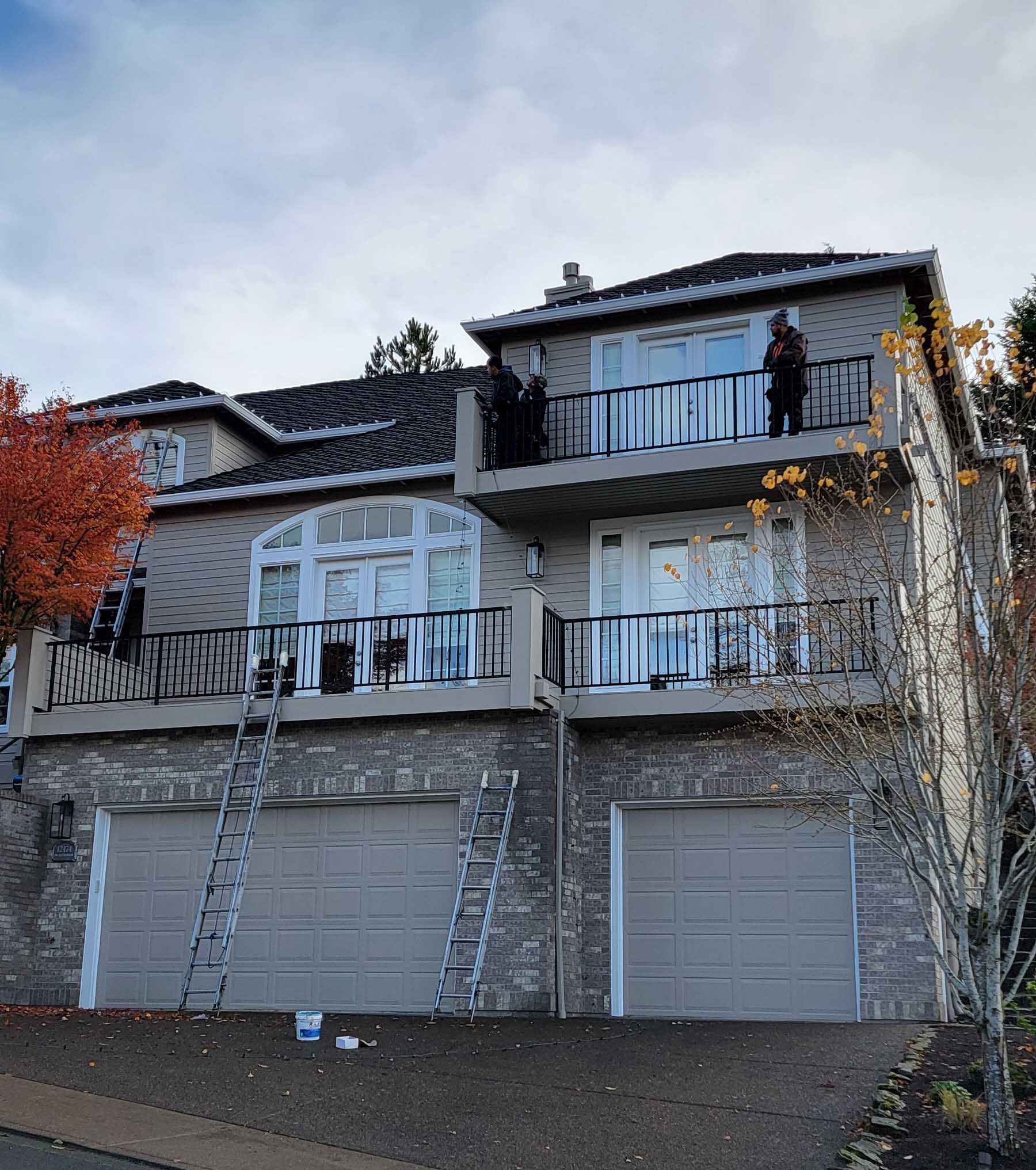 Two-story house with tan siding, brick facade, and garage doors; ladders propped up. People stand on balconies.