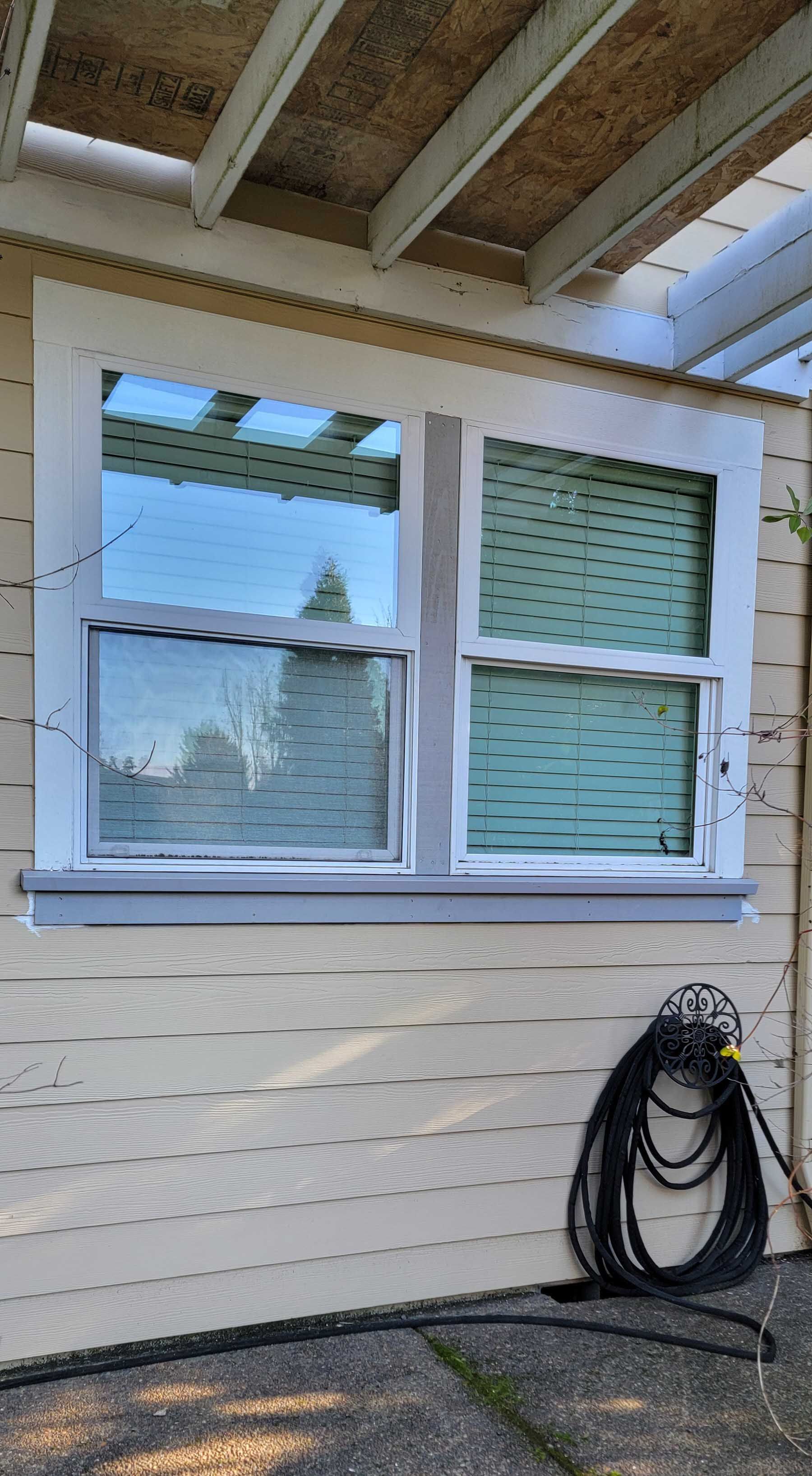 Window with white frame on yellow siding under a pergola; a coiled black hose rests below.