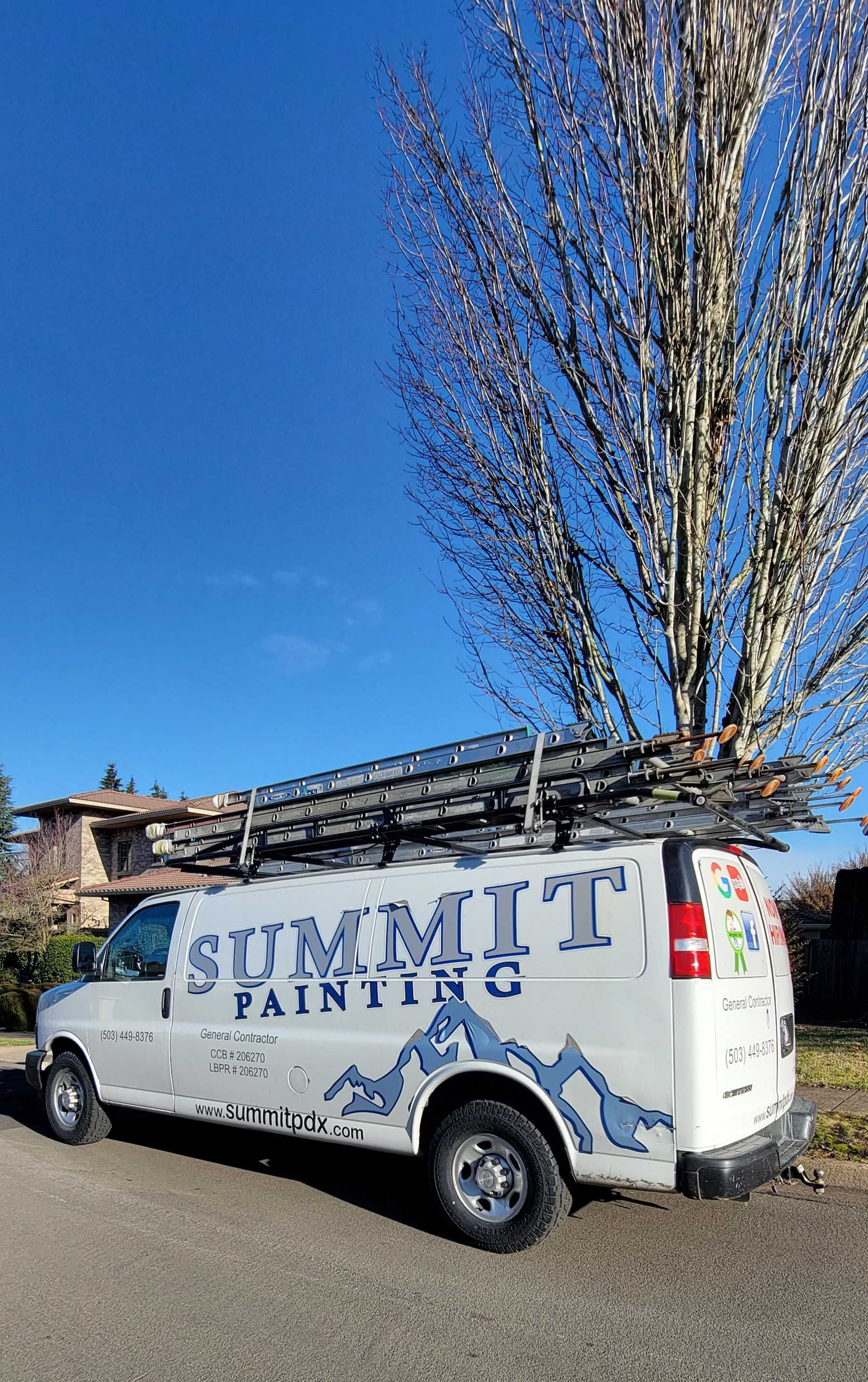 White Summit Painting van with ladders on the roof parked on a road, blue sky, tree in the background.