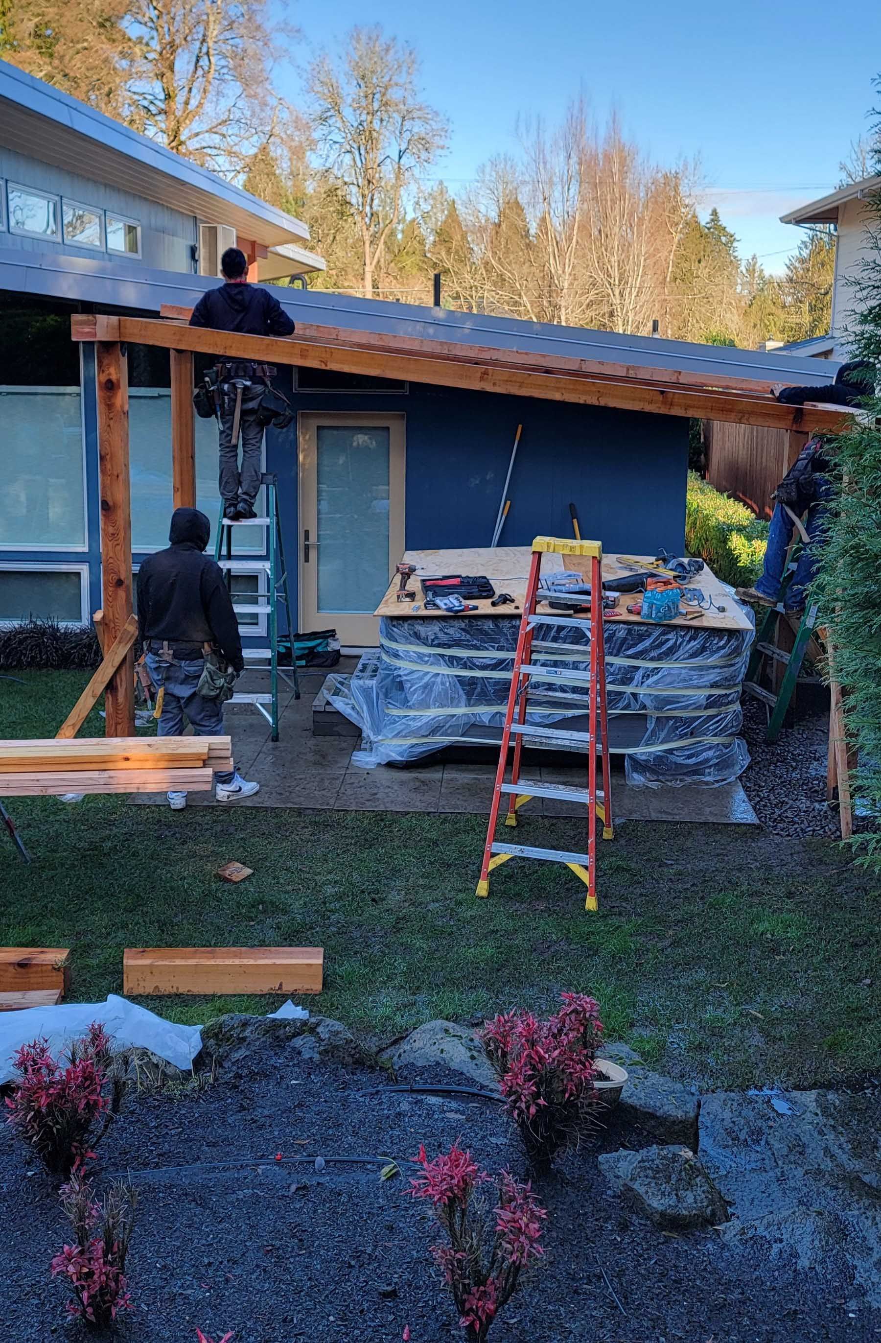 Construction workers building a wooden pergola over a patio next to a blue house.