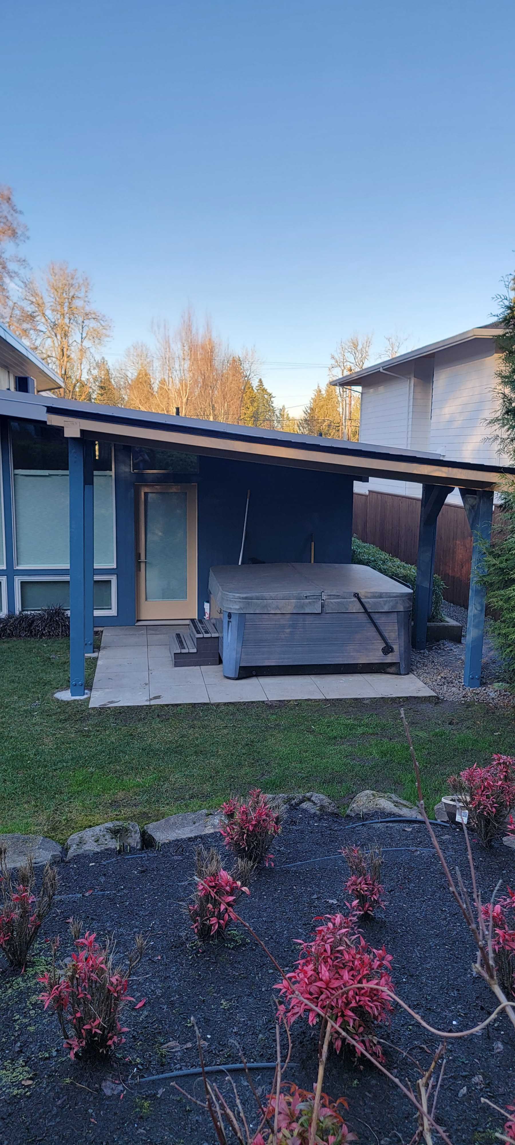 A small house with a dark blue porch and wooden deck; red bushes in the foreground.