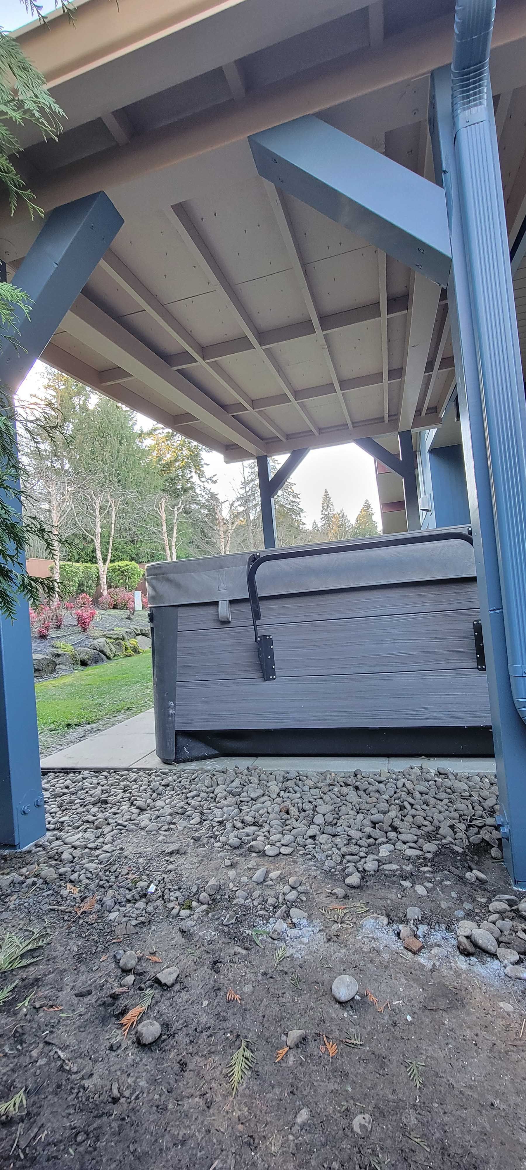 A covered outdoor area with a hot tub. Gray supports and a gravel ground in front.