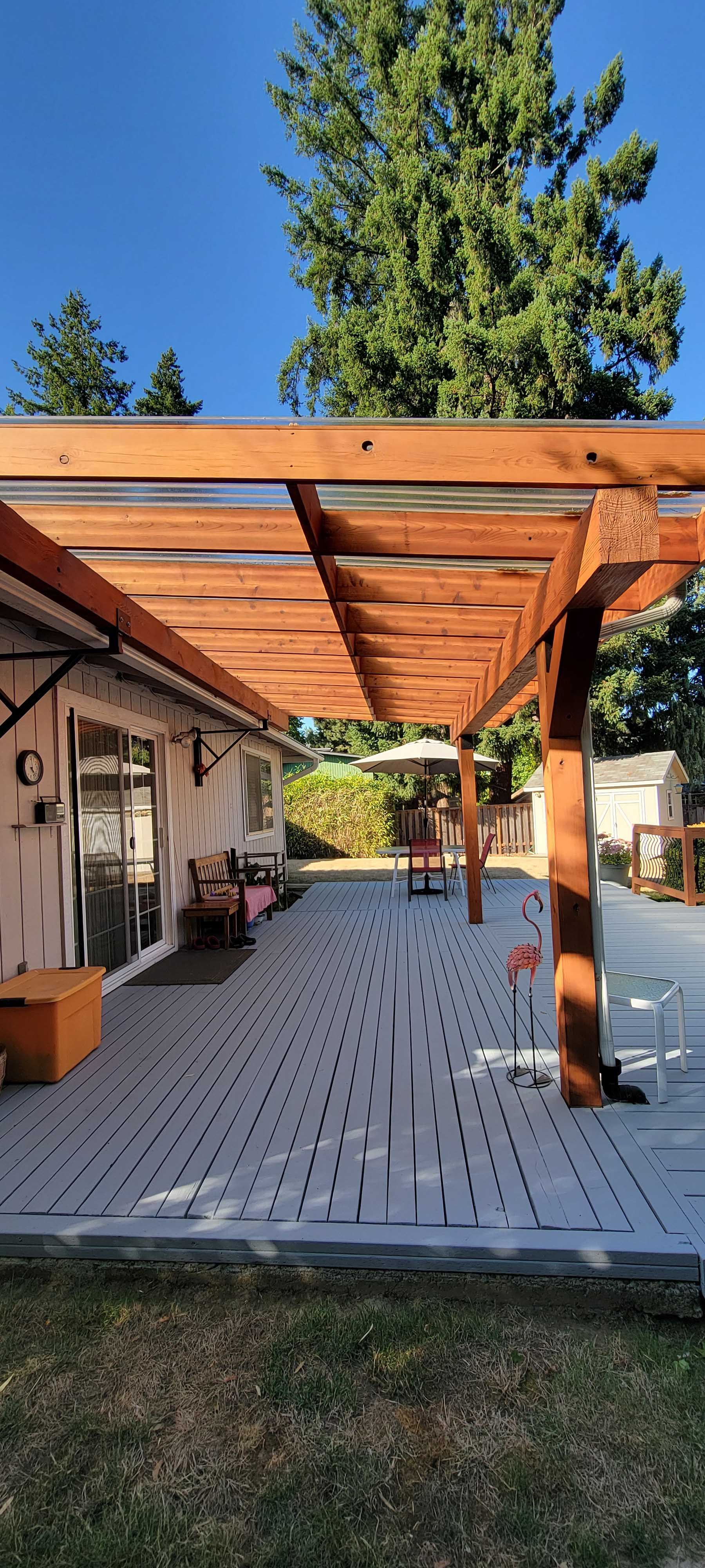 Wooden pergola over a gray patio, with house on the left, trees and blue sky.