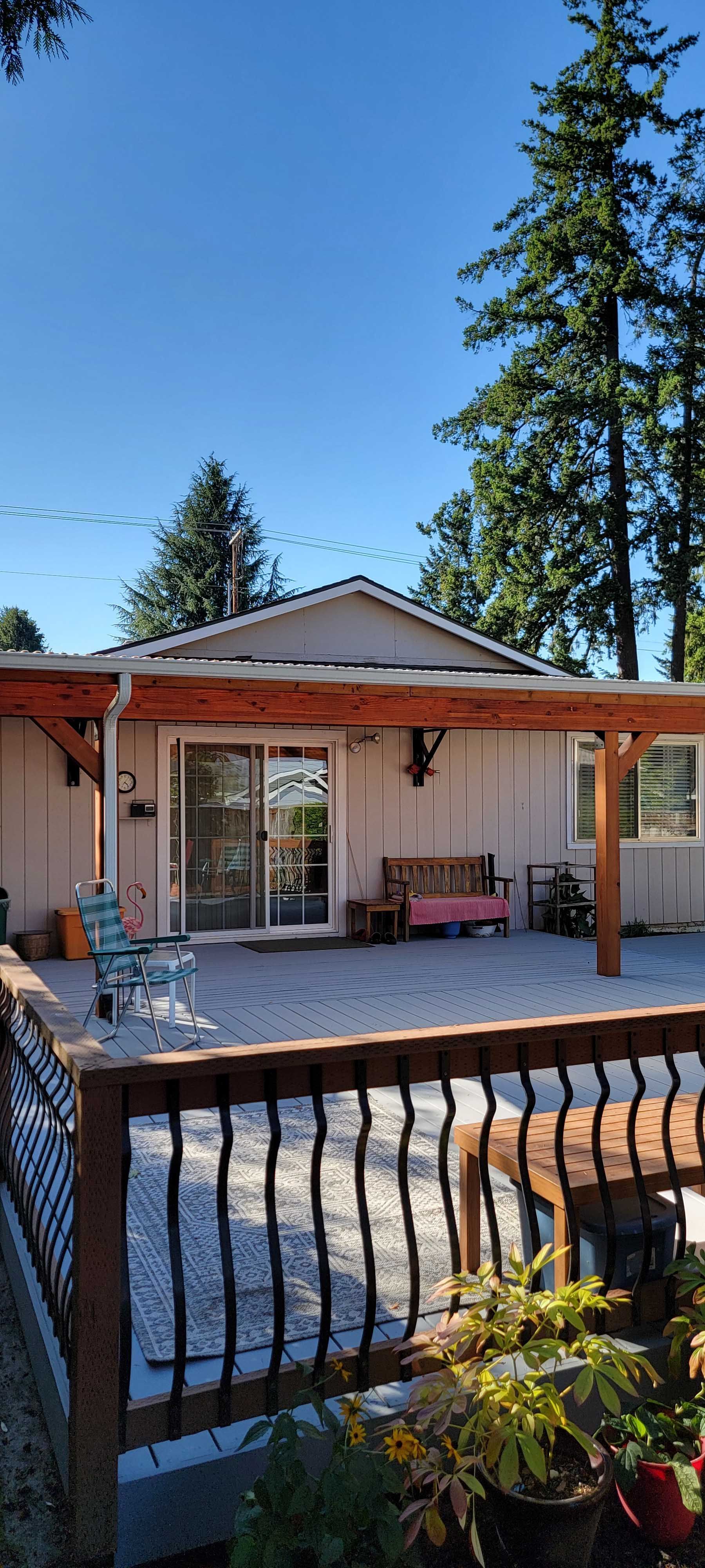 House with a wooden deck, a bench, and a gravel patio, surrounded by trees under a blue sky.