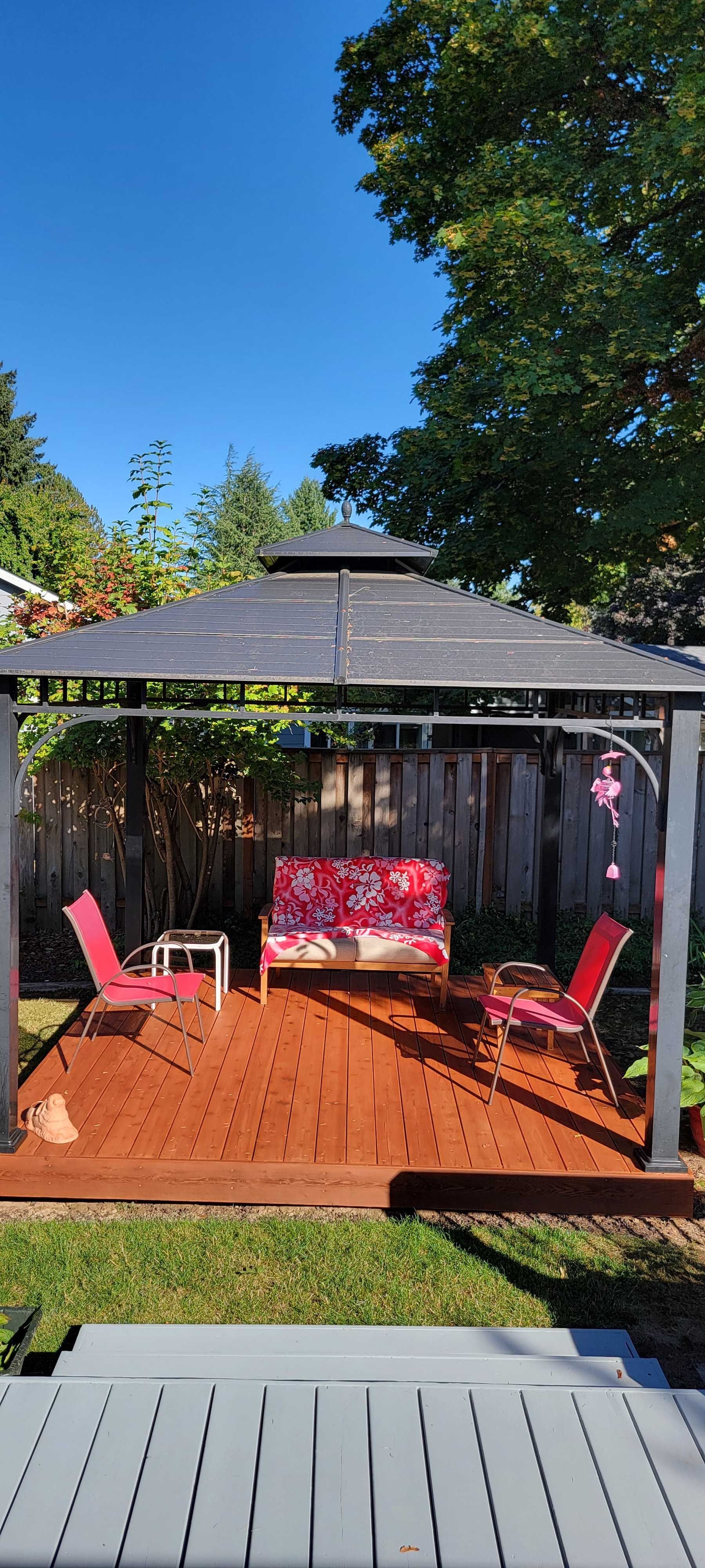 Backyard gazebo with red chairs, a couch, and a small table on a wooden deck. Sunny day.