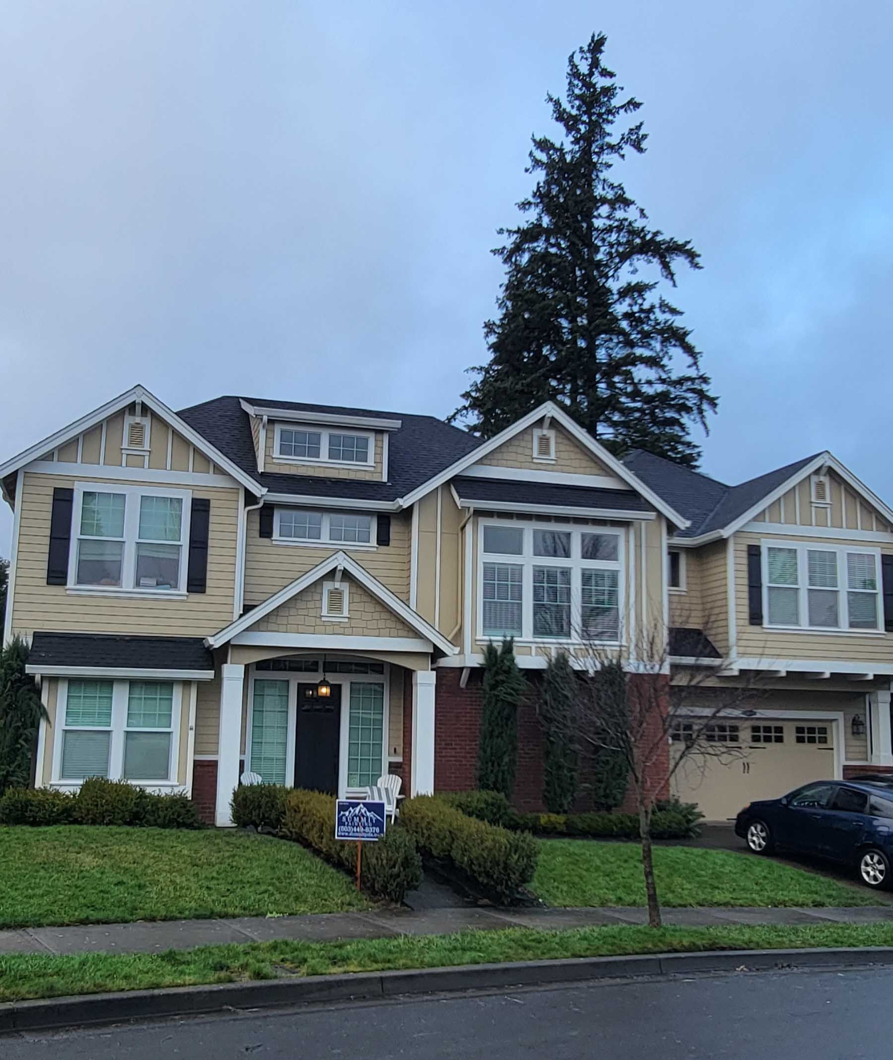 Two-story tan house with black roof and windows, bushes, and a car on a cloudy day.