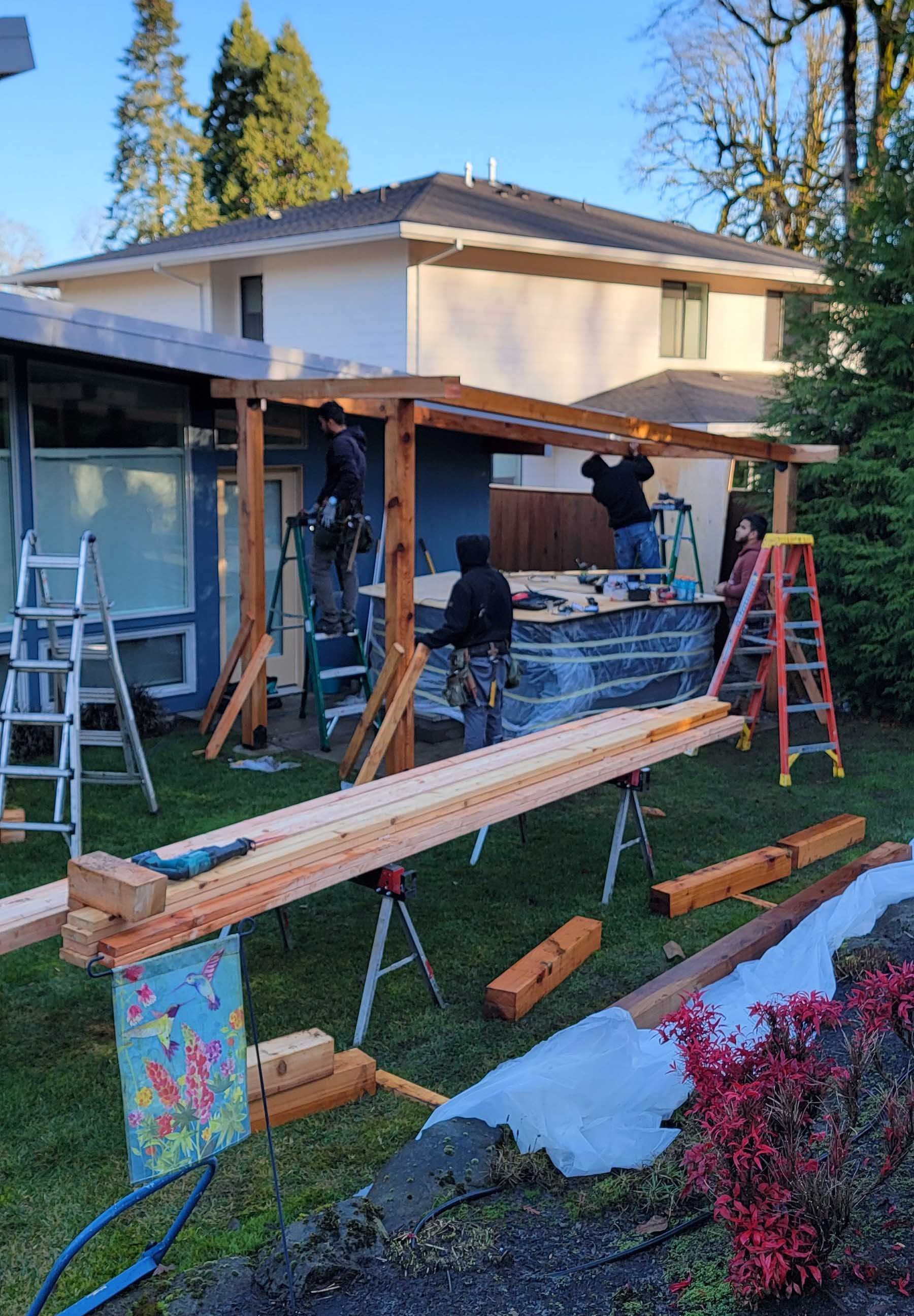 Construction workers building a wooden patio cover on a house. Ladders, tools, and lumber scattered around.