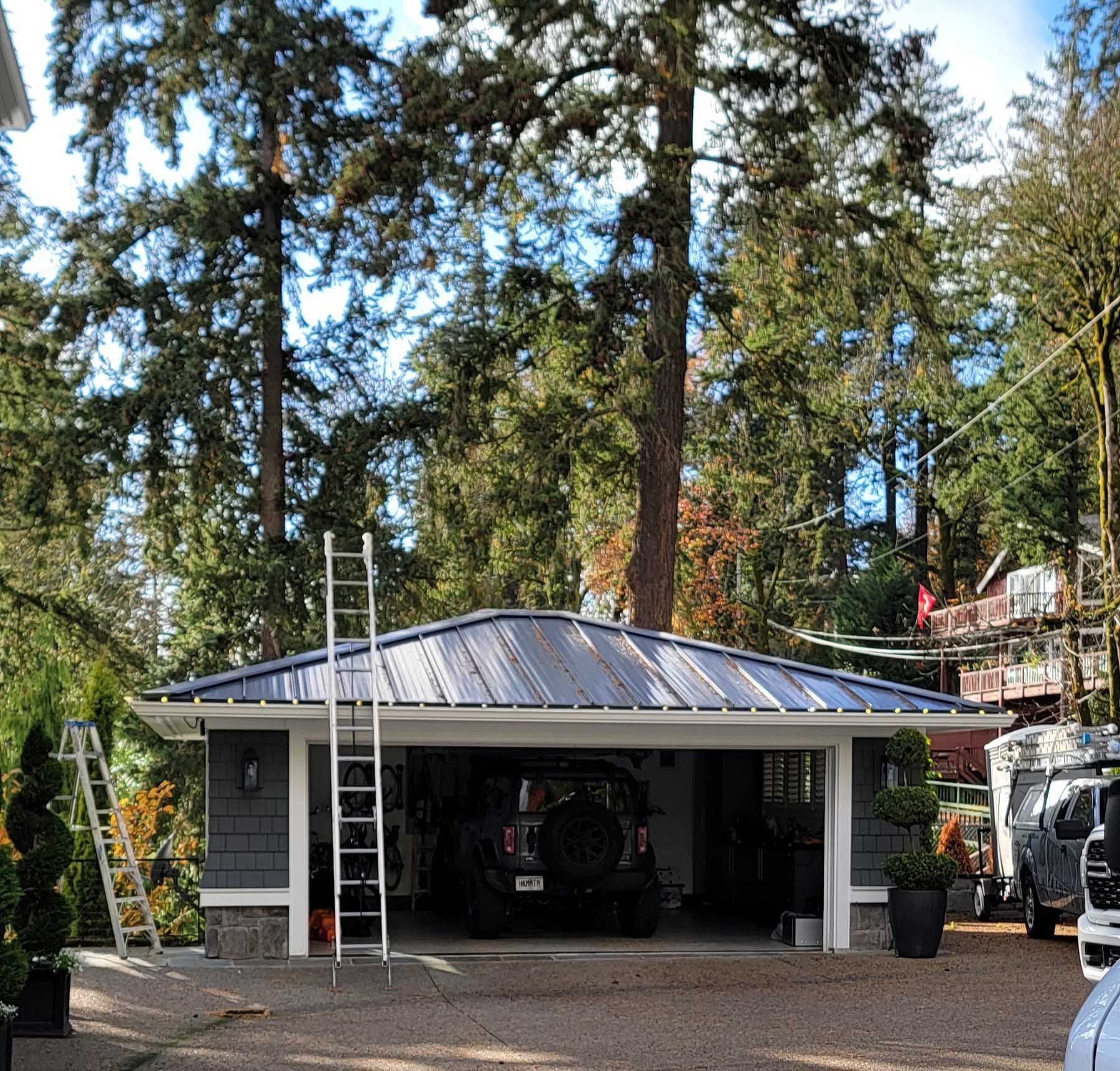 Garage with open doors, gray roof, vehicle inside, surrounded by trees and a ladder.