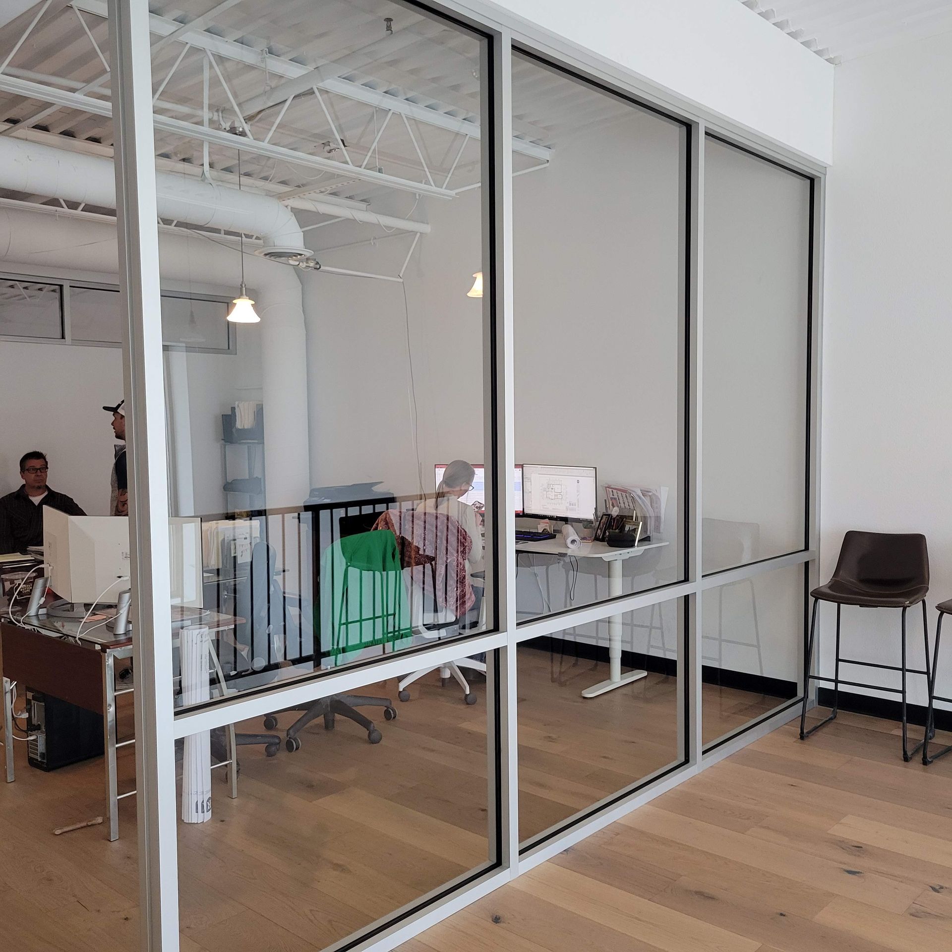 Glass-walled office with people working at desks; light wood floor, white walls and ceiling, two chairs.