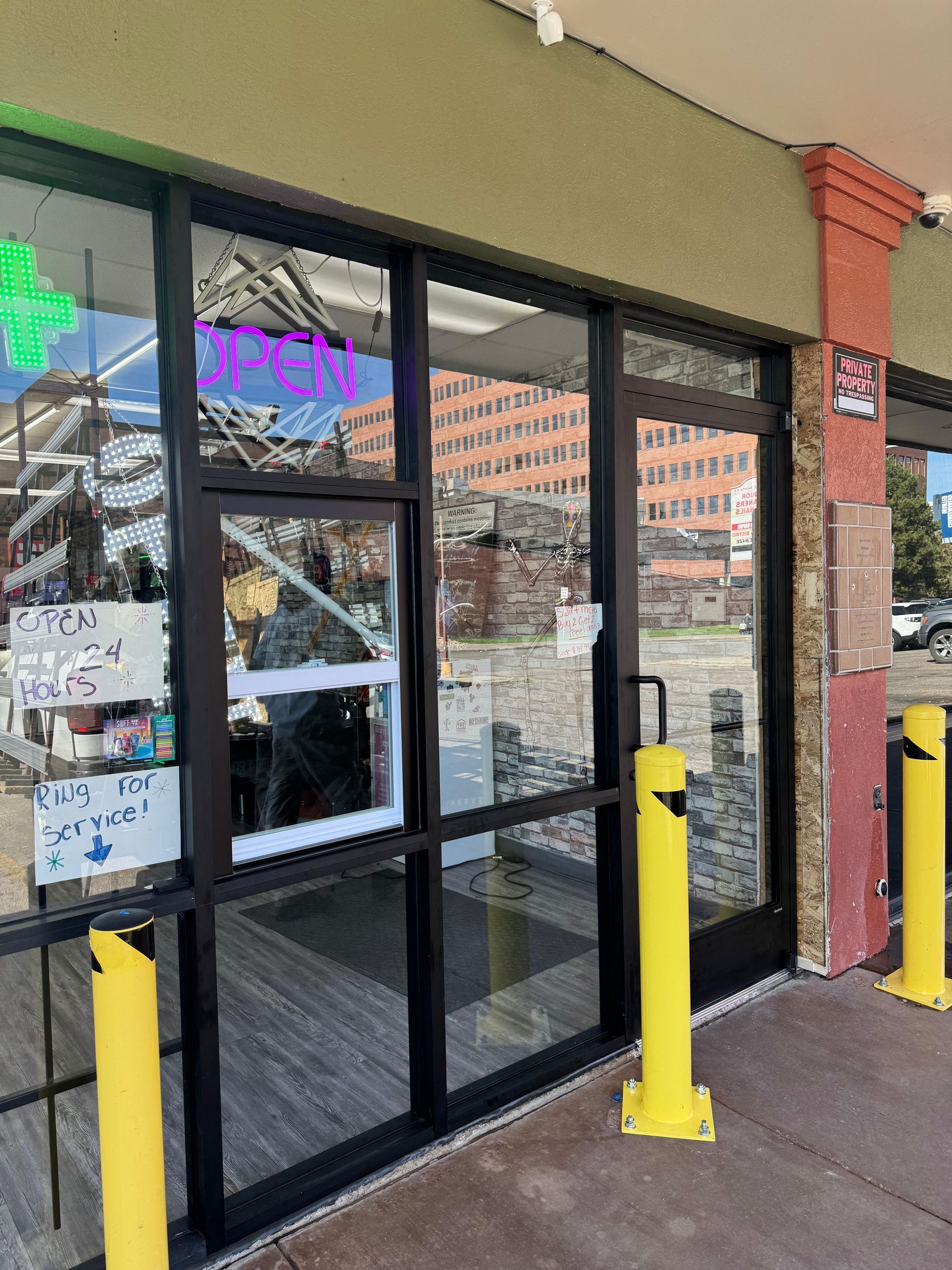 Exterior of a dispensary with glass doors and windows. Yellow safety bollards, a green neon cross, and signs are visible.