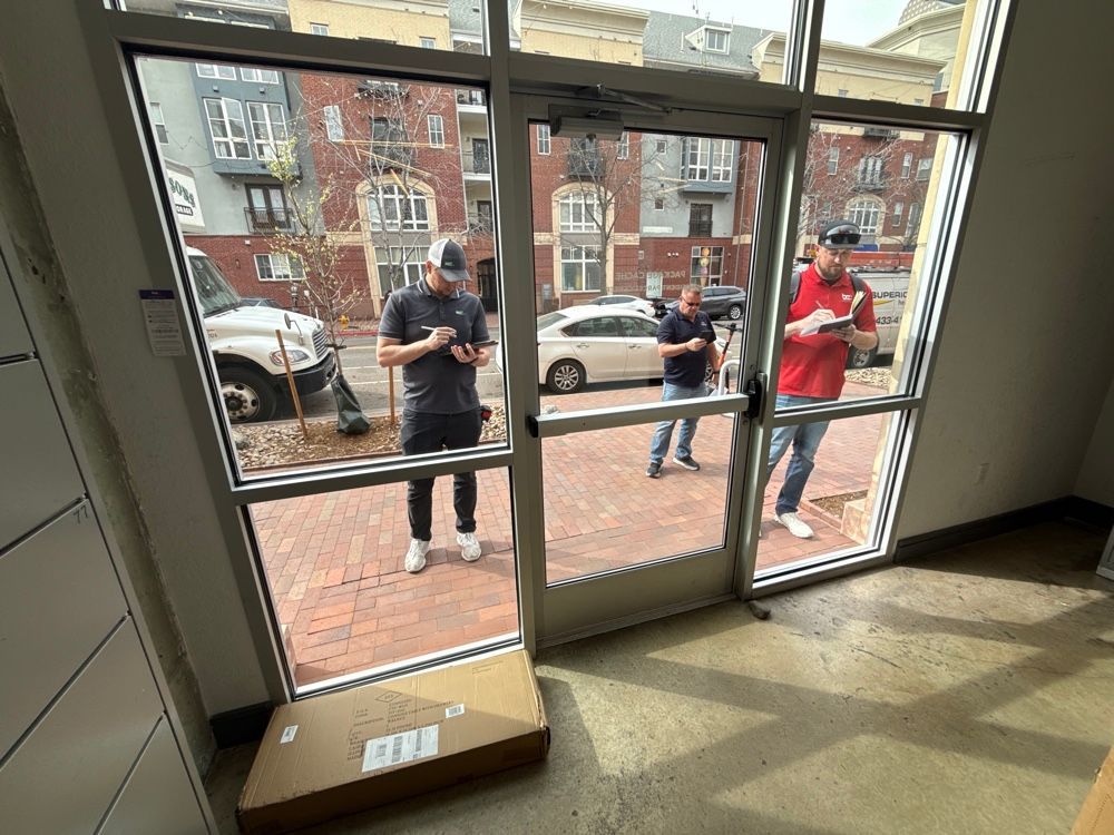Three men in front of a building doorway; one checks phone, two with papers. A package is on the floor.