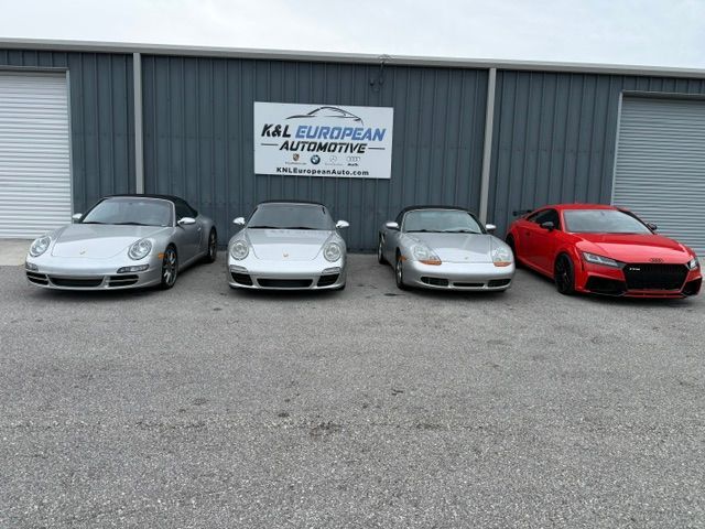 Silver Porsche sports car parked outdoors near other cars under a cloudy sky