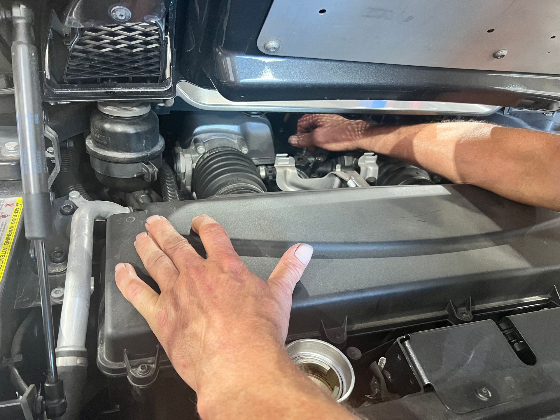Hands working on a car engine, located under a black hood.
