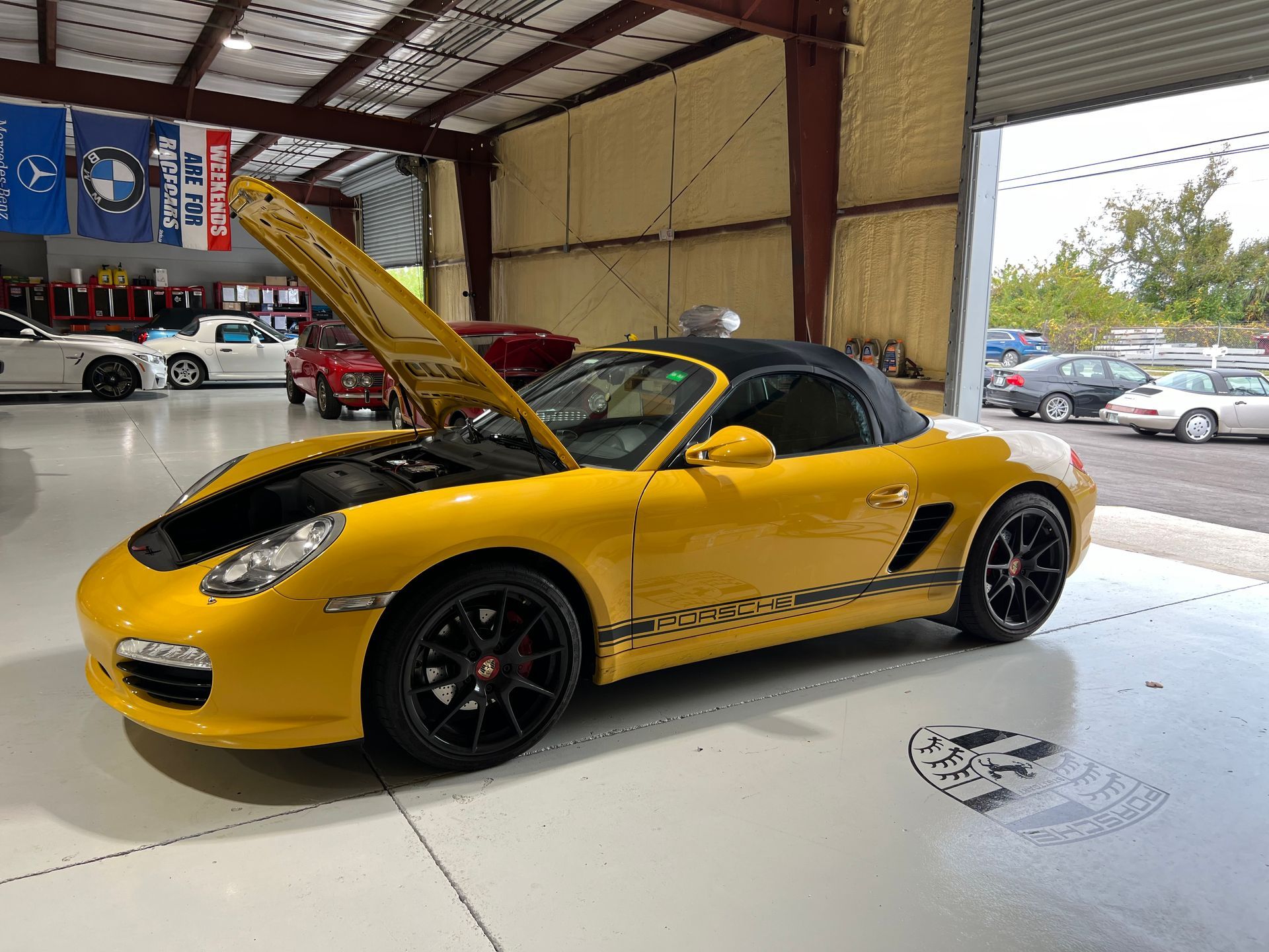 A yellow porsche boxster is parked in a garage with its hood up.