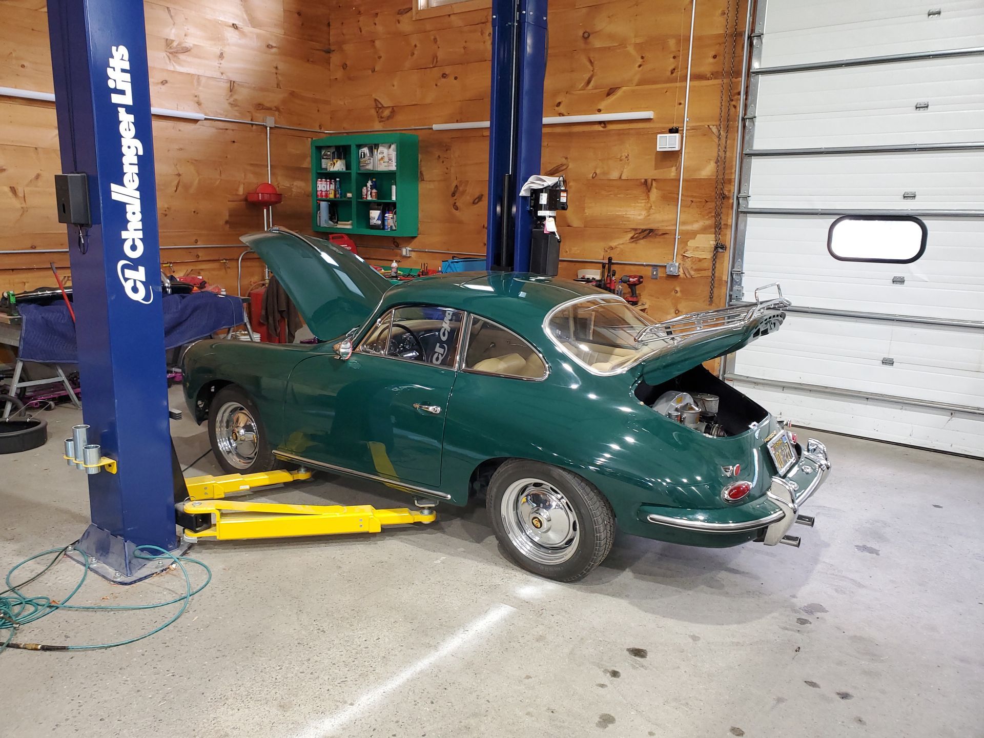 A green car with the hood up is on a lift in a garage.