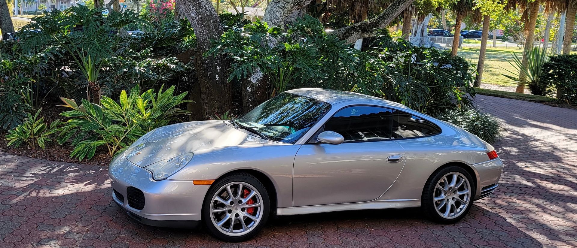 A silver porsche 911 is parked in a driveway next to trees.