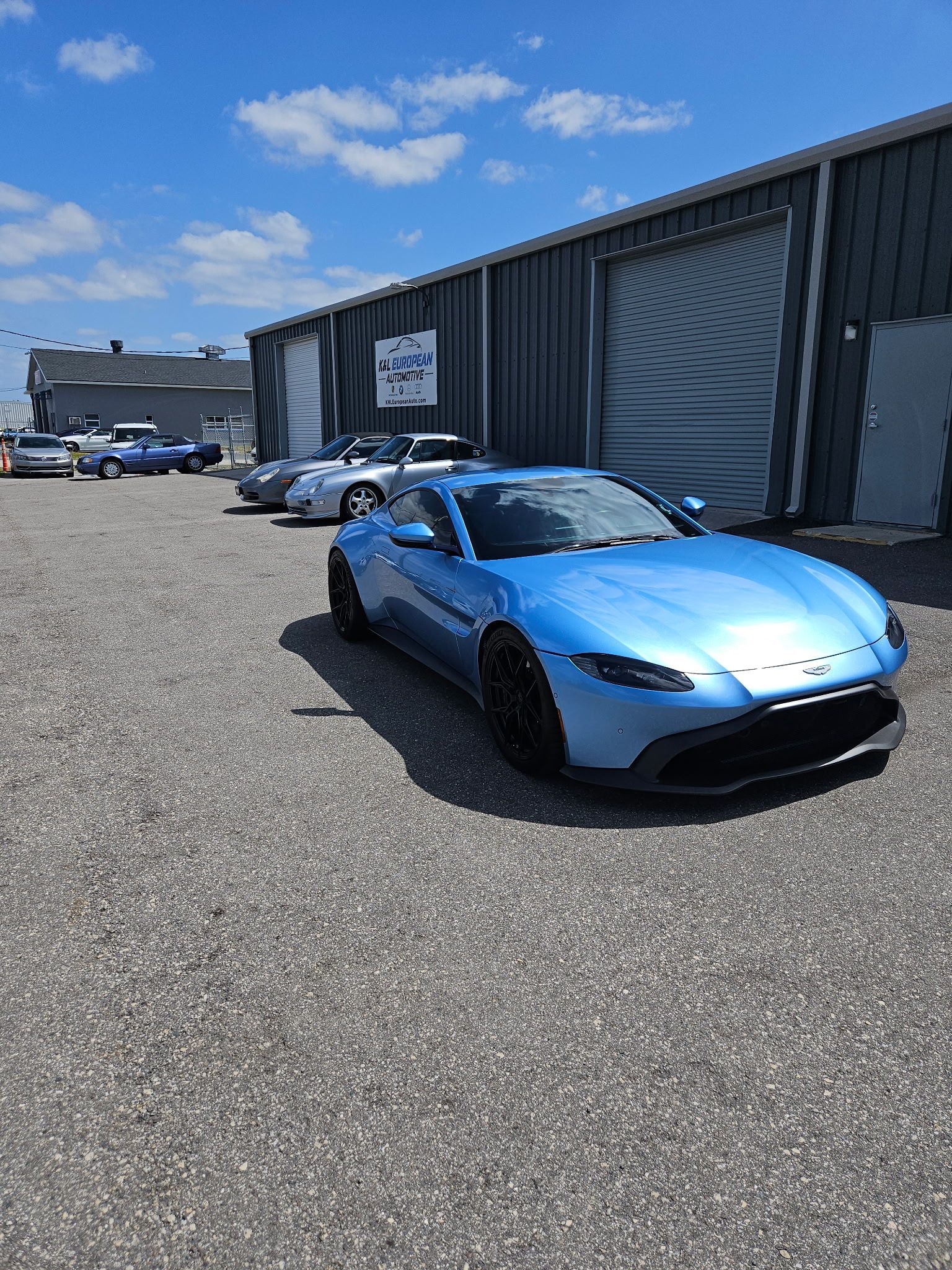 A blue sports car is parked in a gravel lot in front of a building.
