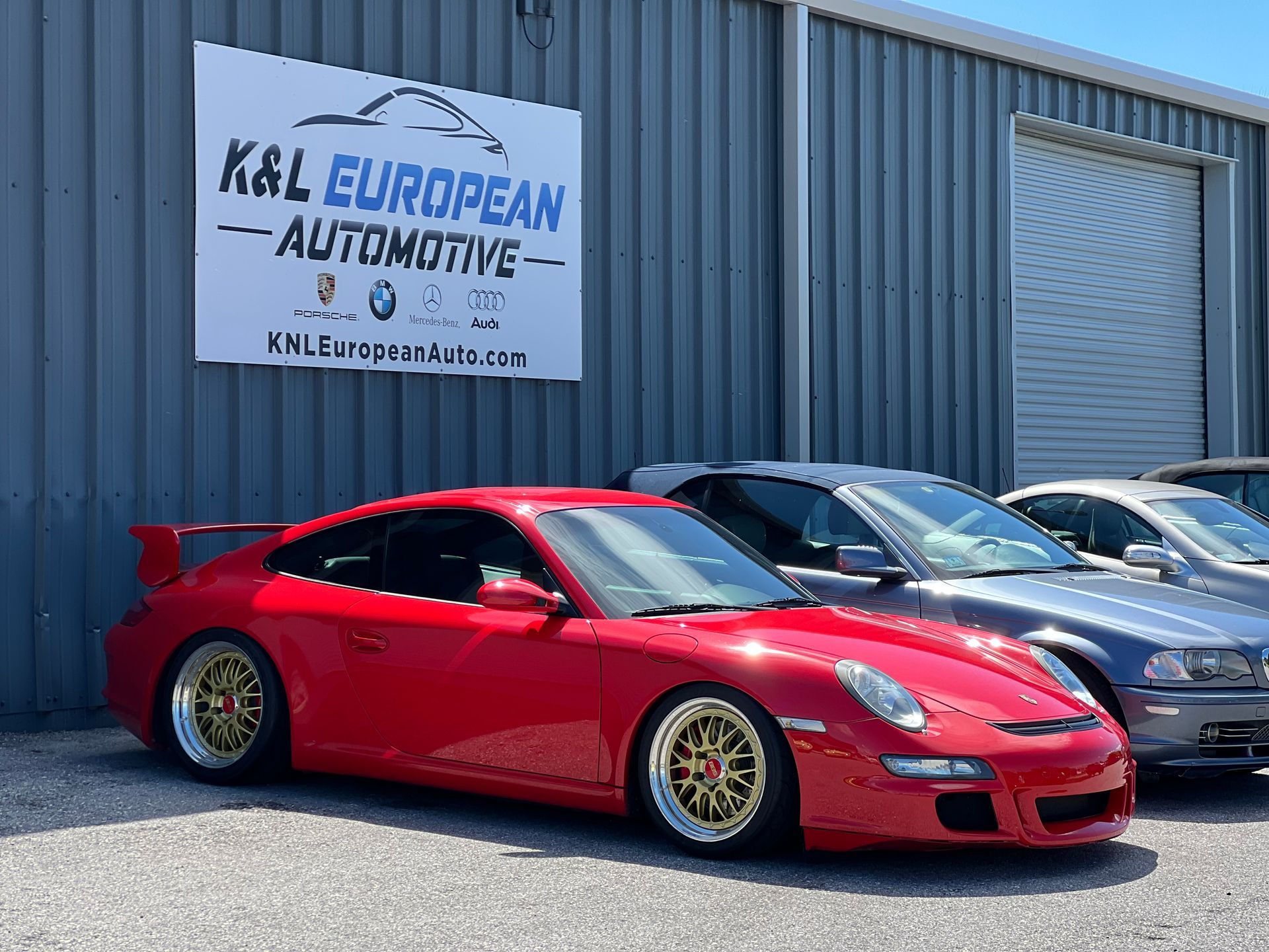 Red Porsche sports car parked outside K&L European Automotive shop.